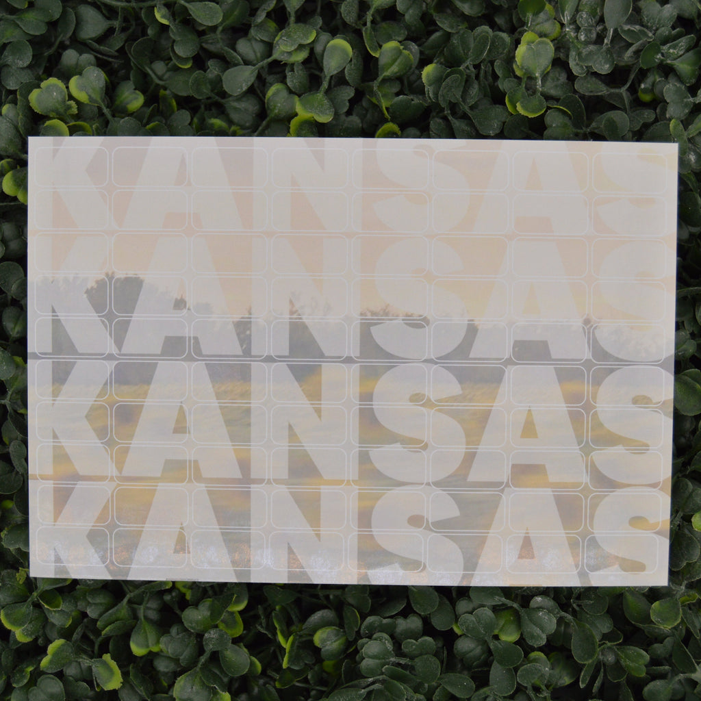 A square wall art print featuring the word 'KANSAS' repeated vertically in large white letters over a soft sunset landscape, displayed against a green leafy hedge background.
