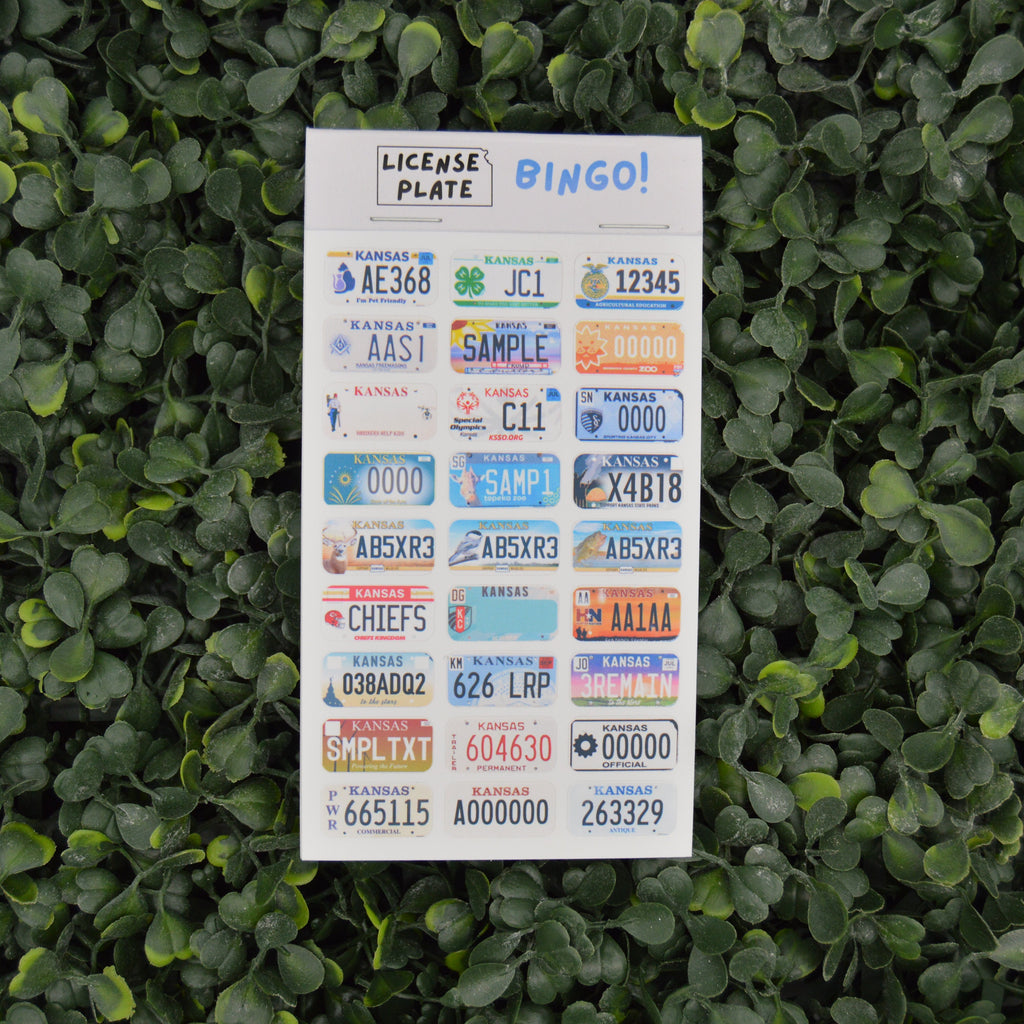 A sheet of novelty license plate bingo stickers displayed against a background of green ivy leaves. The white sticker sheet features various colorful sample license plates with placeholder text and numbers, topped with 'LICENSE PLATE BINGO!' in blue and black lettering.