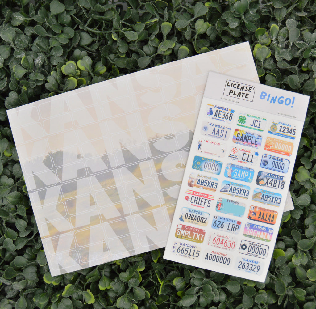 A sheet of colorful novelty license plate stickers labeled 'LICENSE PLATE BINGO!' is placed on top of a white envelope or card, both resting on a bed of green leaves.