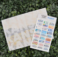 A sheet of colorful novelty license plate stickers labeled 'LICENSE PLATE BINGO!' is placed on top of a white envelope or card, both resting on a bed of green leaves.