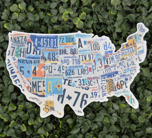 A decorative silhouette map of the United States made from colorful vintage license plates, displayed against a dense green leafy hedge background.