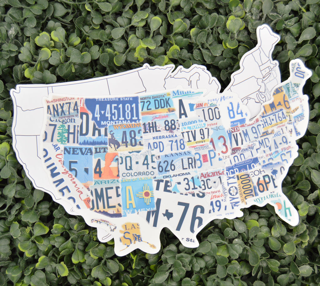A die-cut map of the United States made from colorful vintage license plates, displayed on a background of green artificial foliage.