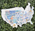 A die-cut map of the United States made from colorful vintage license plates, displayed on a background of green artificial foliage.