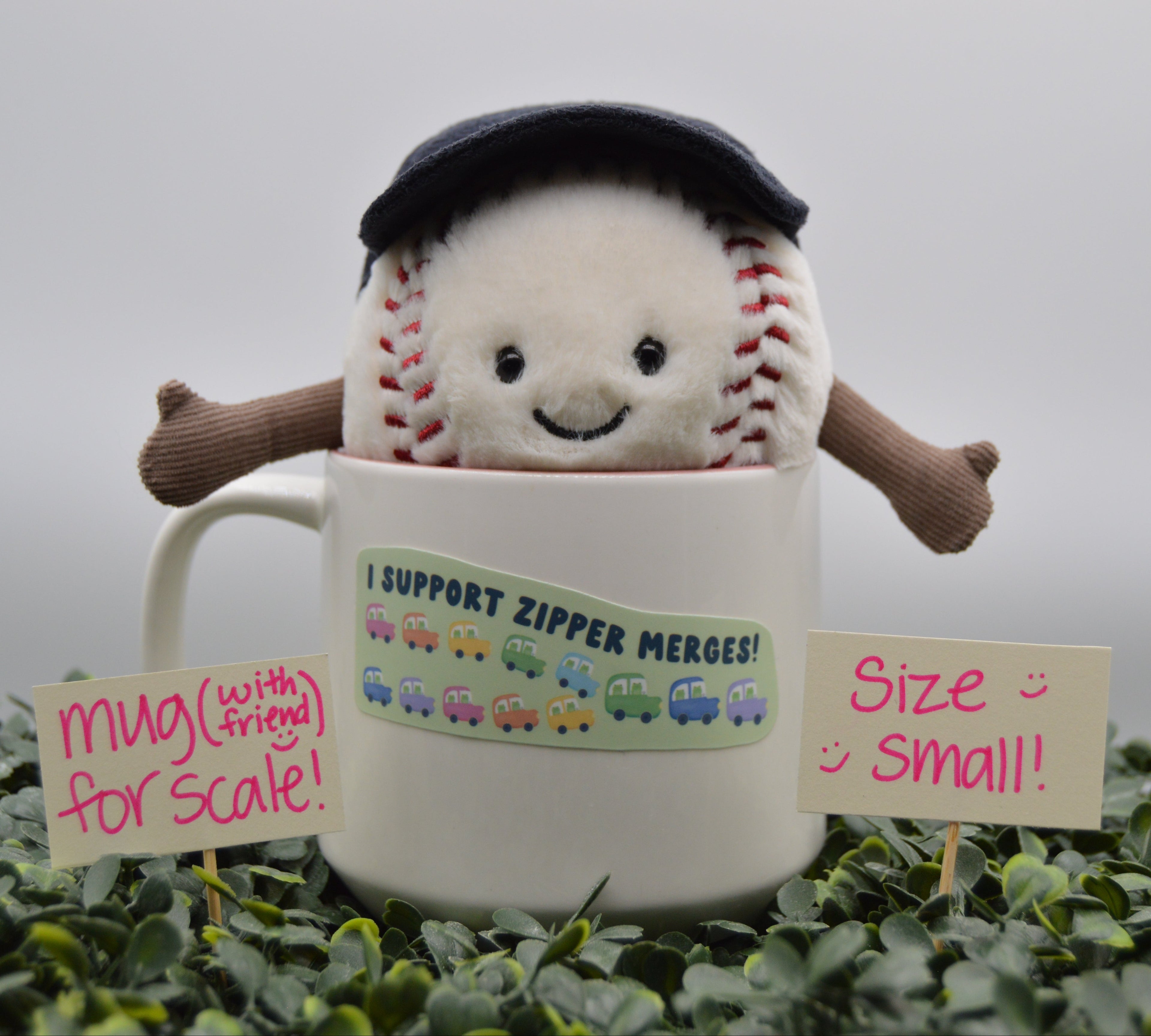 A plush baseball character wearing a black cap and brown arms pops out of a white ceramic mug. The mug has a sticker that reads 'I SUPPORT ZIPPER MERGES!'.