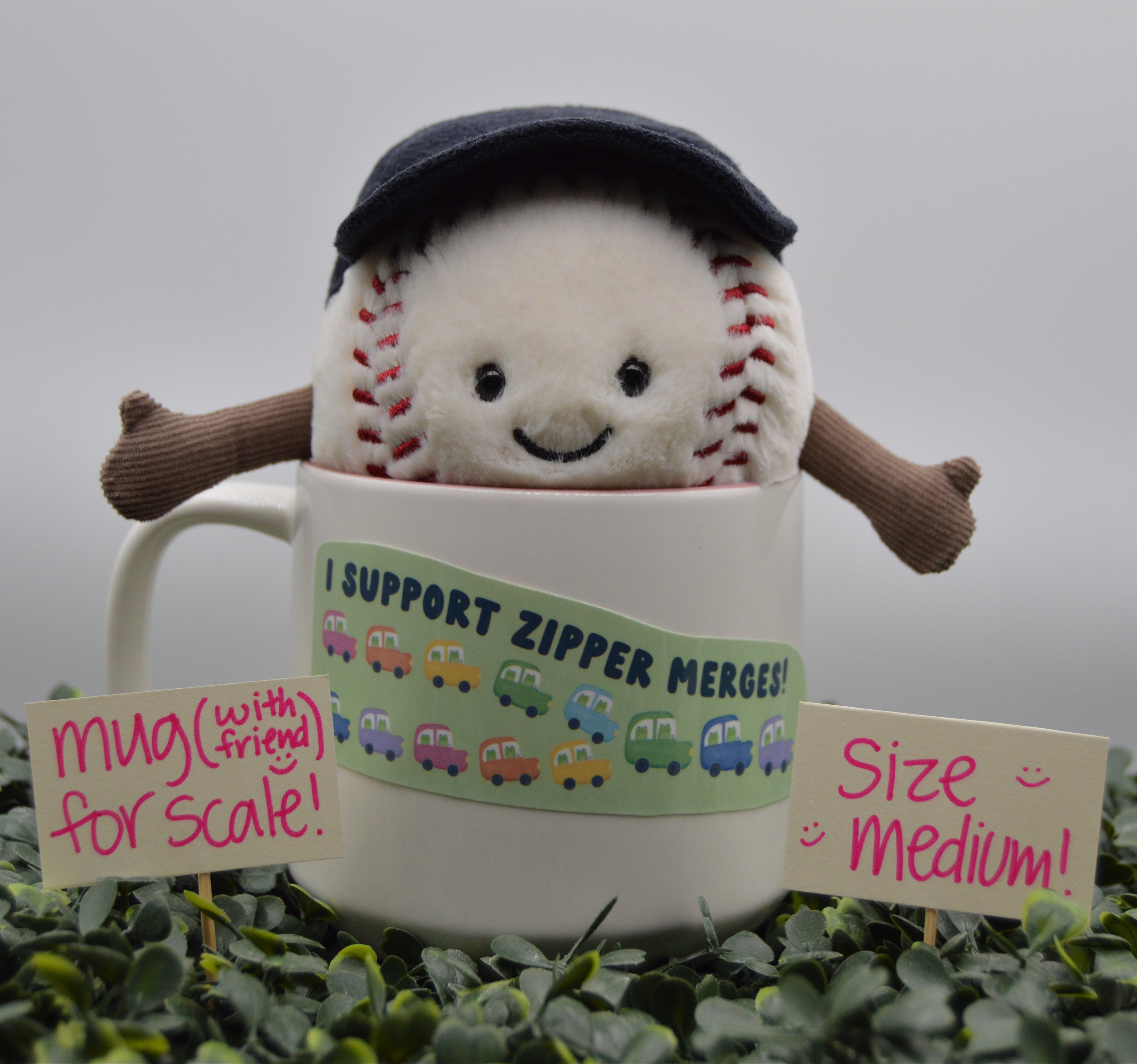 A plush baseball character wearing a dark navy cap and brown arms, emerging from a white ceramic mug. The mug has a green band reading 'I SUPPORT ZIPPER MERGE!' with small colorful cars.