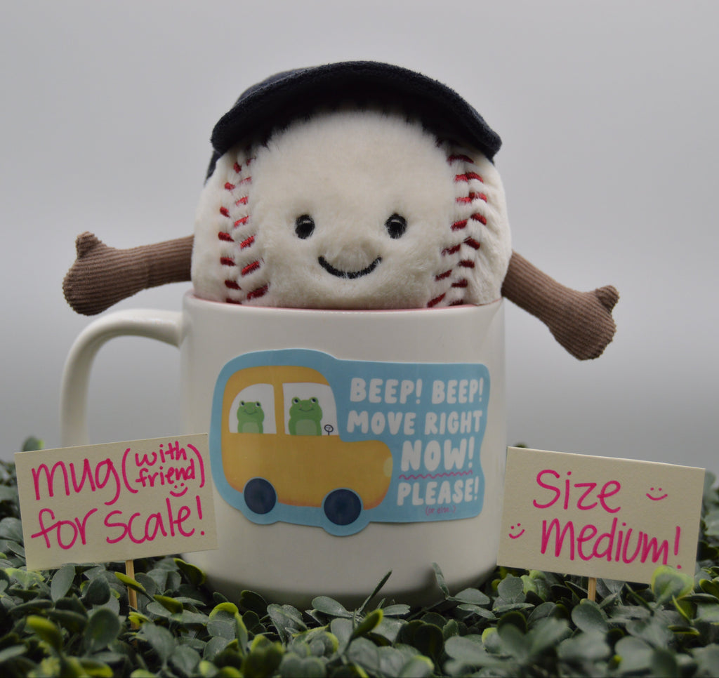 A plush baseball character wearing a black beret and brown arms sits inside a white ceramic mug with a blue and yellow bus graphic and the text 'BEEP! BEEP! MOVE RIGHT NOW! PLEASE!'. The mug is surrounded by green foliage with two small signs reading 'mug (with friend) for scale!' and 'Size Medium!'.