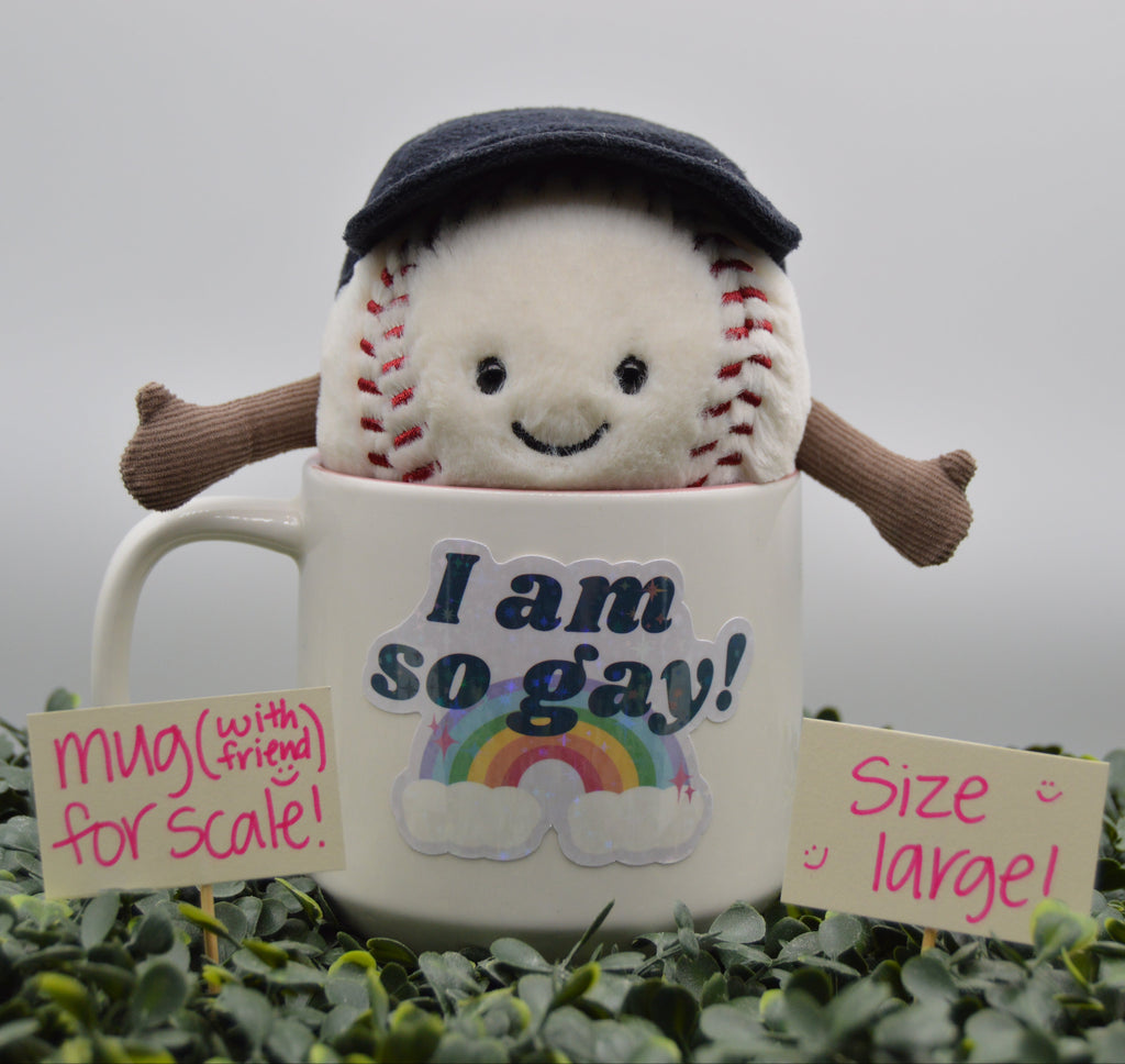 A plush baseball toy wearing a dark navy cap and brown arms, emerging from a white ceramic mug printed with the text 'I am so gay!' and a rainbow. The setup is surrounded by green foliage, with two pink handwritten notes reading 'mug (with friend for scale!)' and 'size large!'.