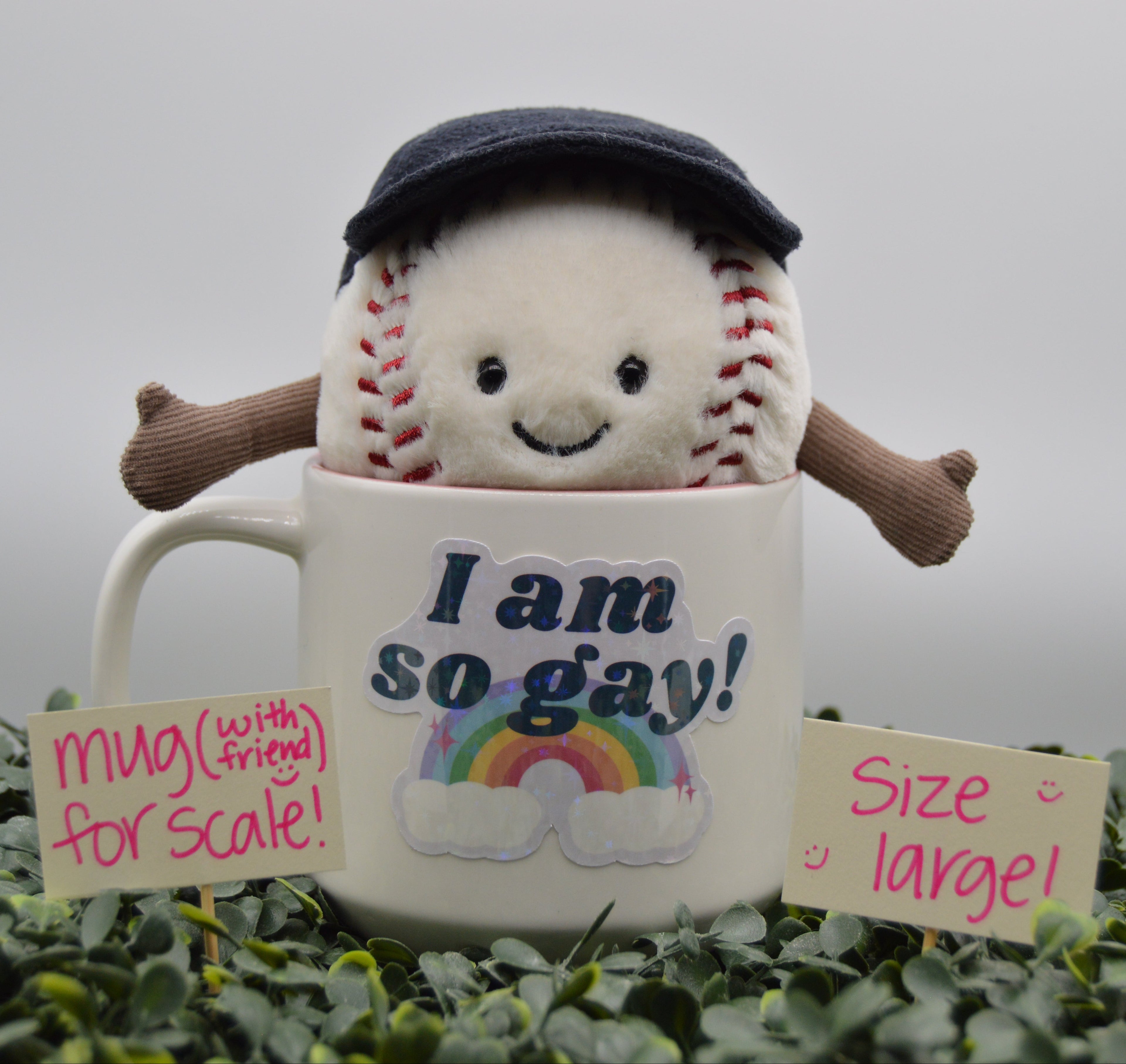 A plush baseball toy wearing a dark navy cap and brown arms, emerging from a white ceramic mug printed with the text 'I am so gay!' and a rainbow. The setup is surrounded by green foliage, with two pink handwritten notes reading 'mug (with friend for scale!)' and 'size large!'.