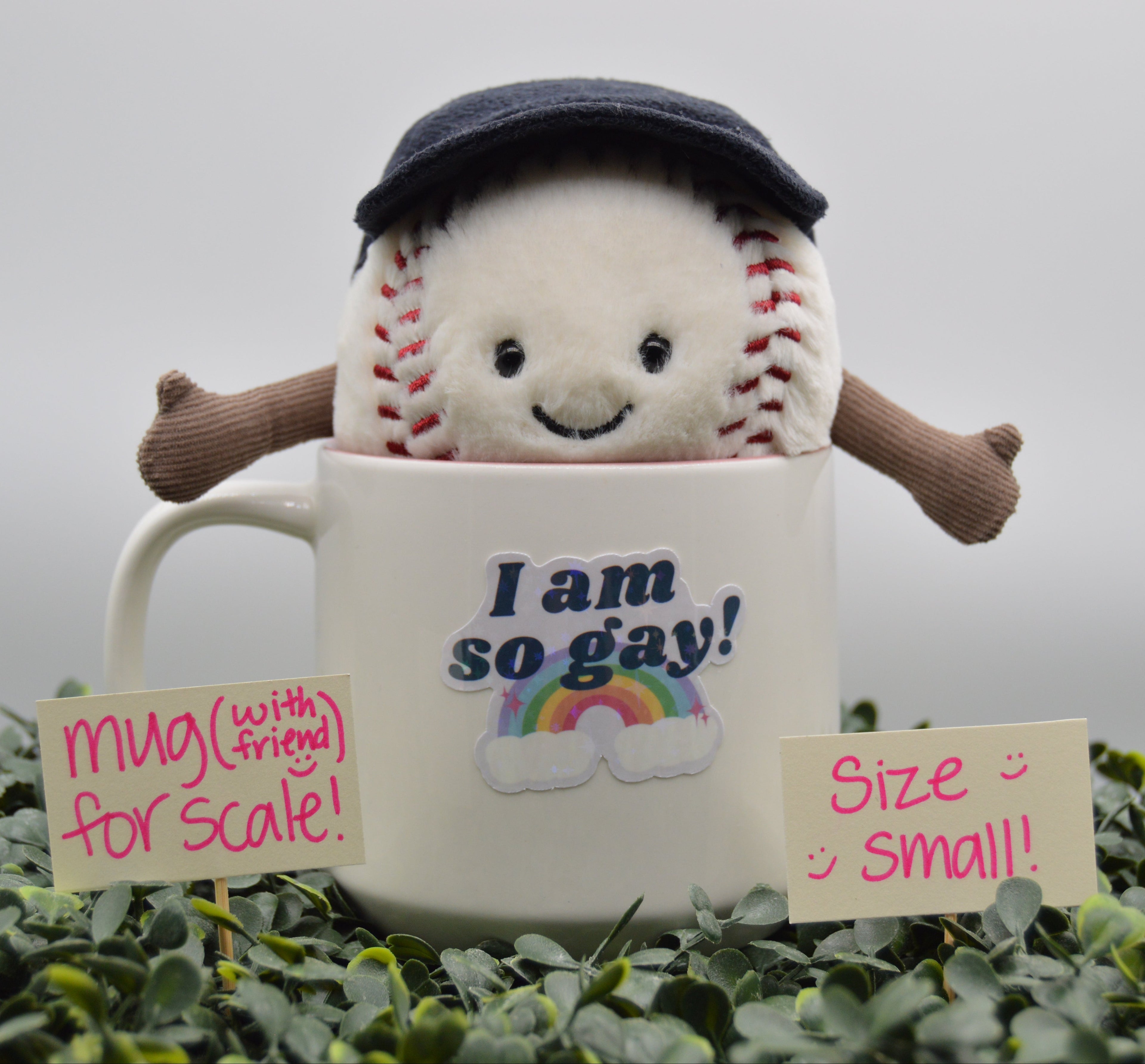 A small plush baseball toy wearing a dark navy cap and brown arms, emerging from a white ceramic mug printed with the phrase 'I am so gay!' and a rainbow. The setup is surrounded by green foliage, with two small handwritten signs reading 'mug (with friend) for scale!' and 'Size: small!!'.