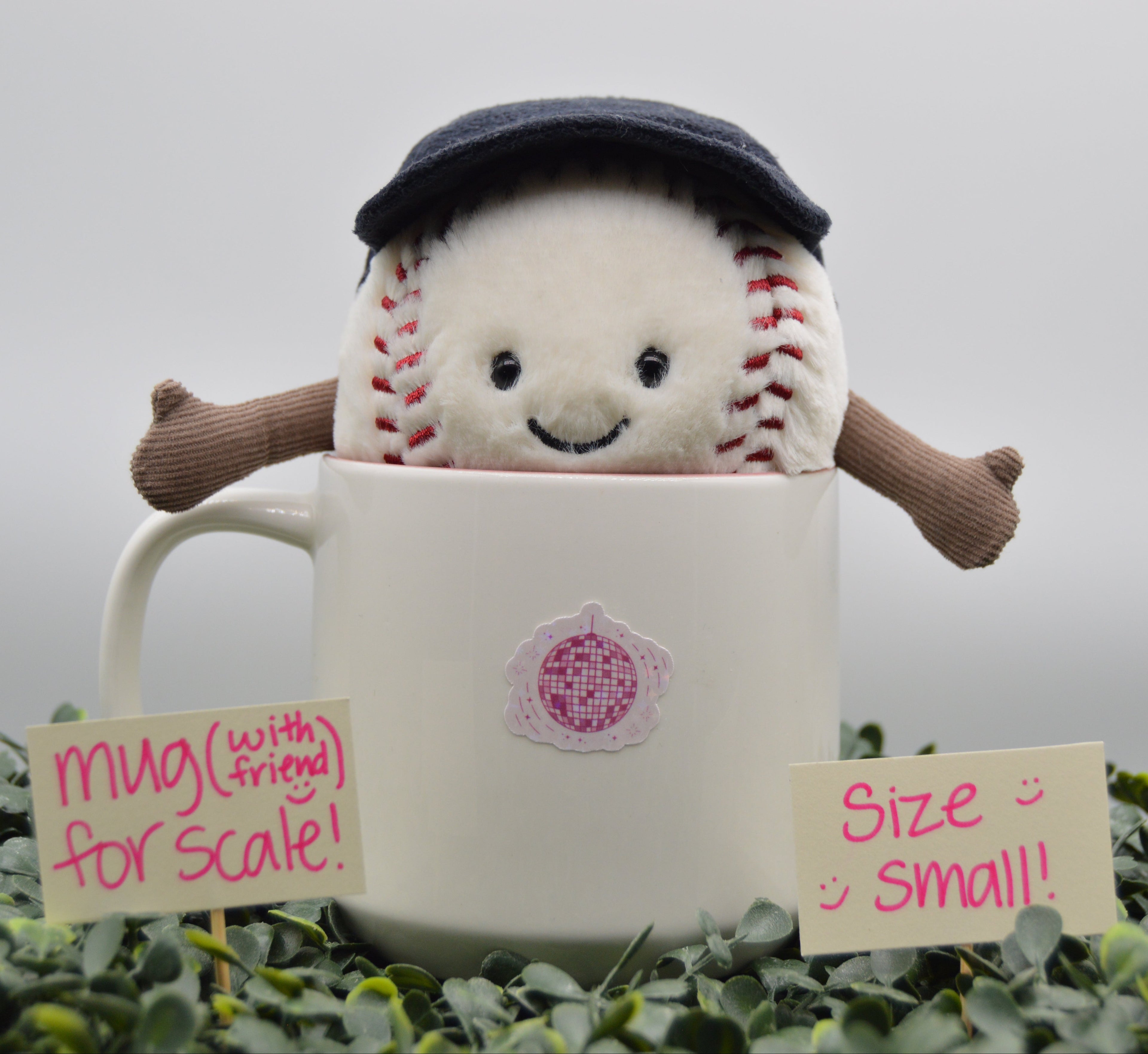 A plush baseball toy peeking out of a white ceramic mug, with handwritten notes in front reading 'mug (with friend) for scale!' and 'Size small!!'. The background is a soft gray gradient with green foliage at the bottom.