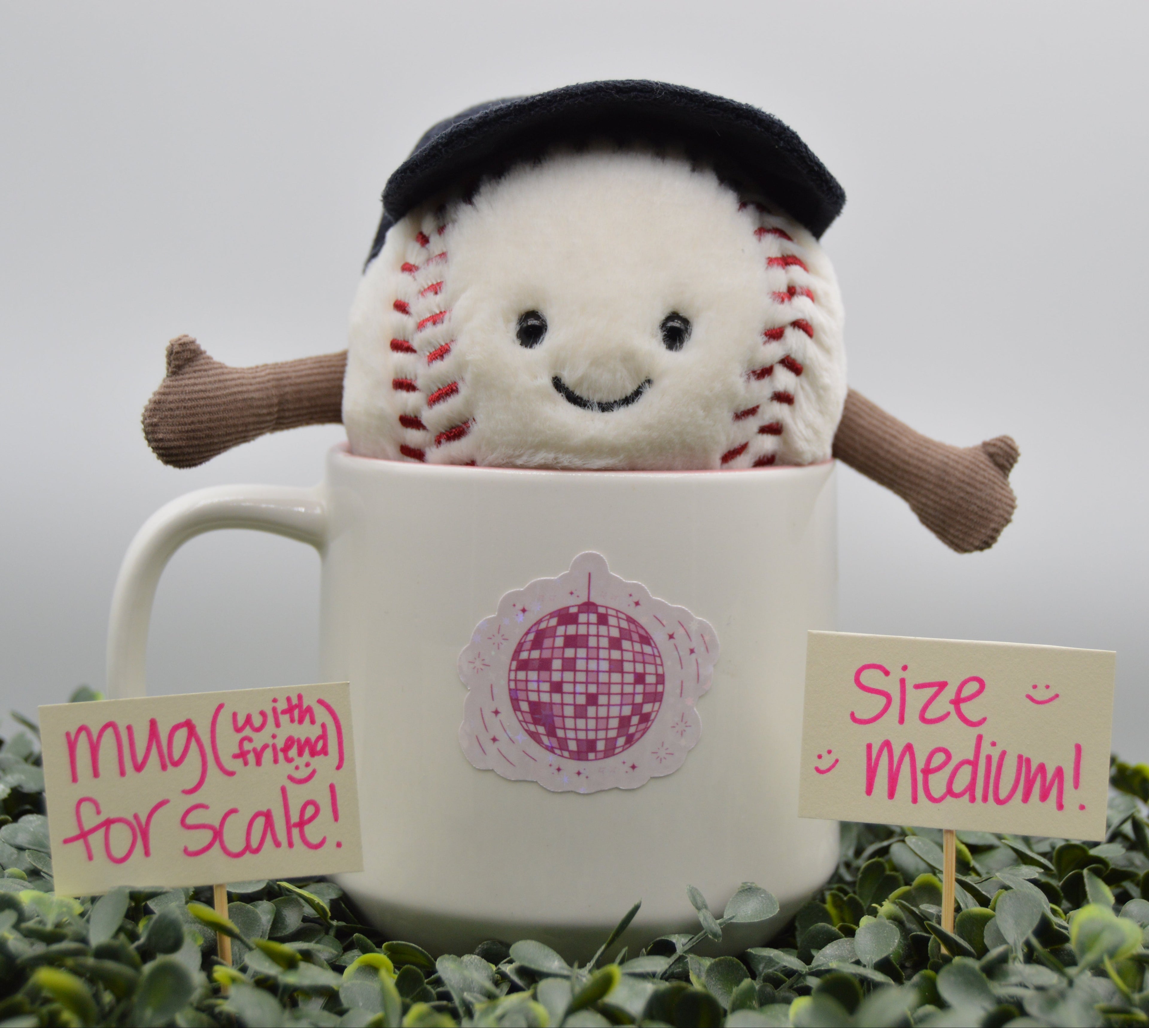 A plush baseball character wearing a black beret sits inside a white ceramic mug, surrounded by green foliage with handwritten scale and size signs in front.