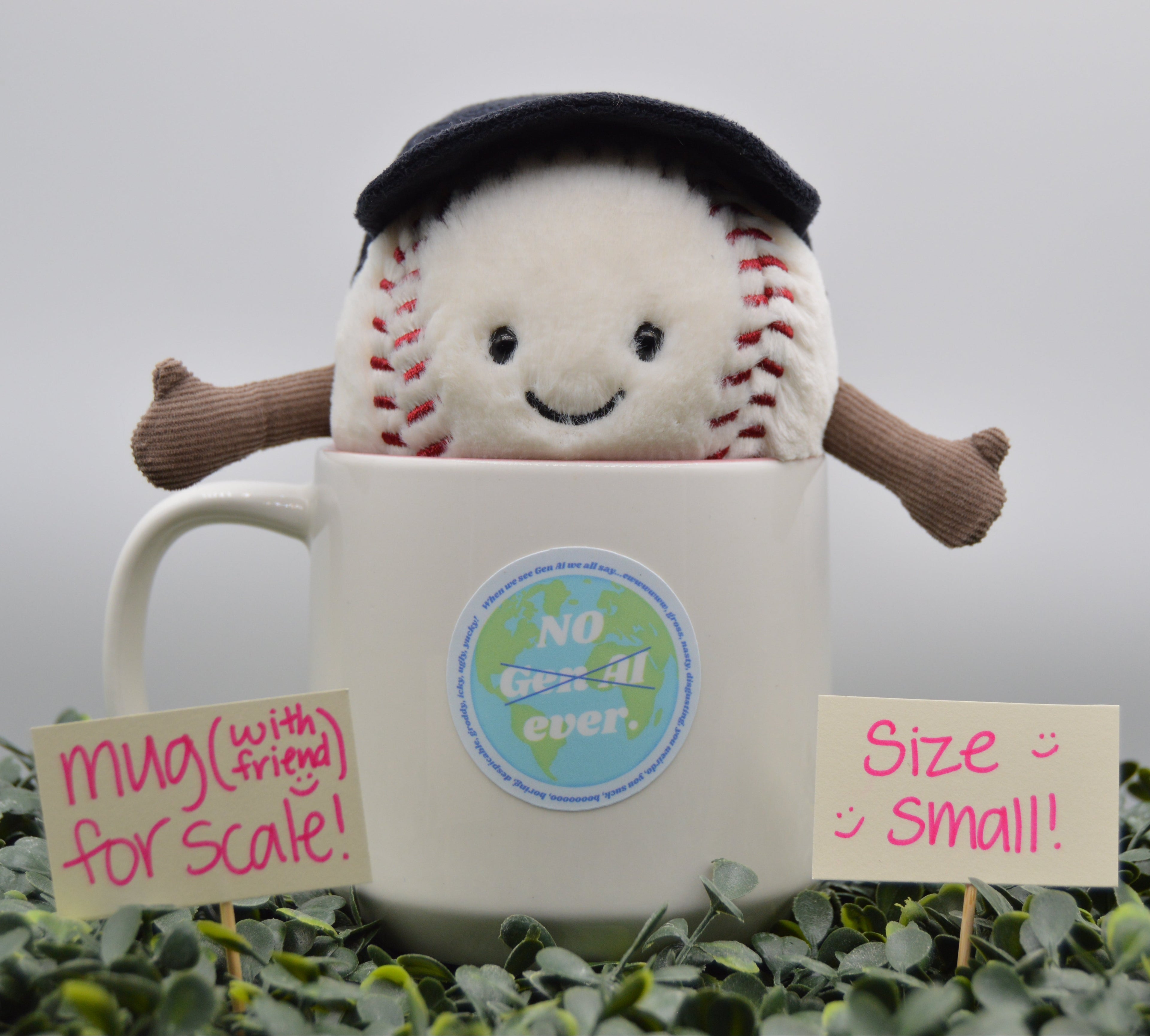 A plush baseball character toy with a black beret and brown arms sits inside a white ceramic mug, surrounded by green foliage with handwritten scale signs in the foreground.