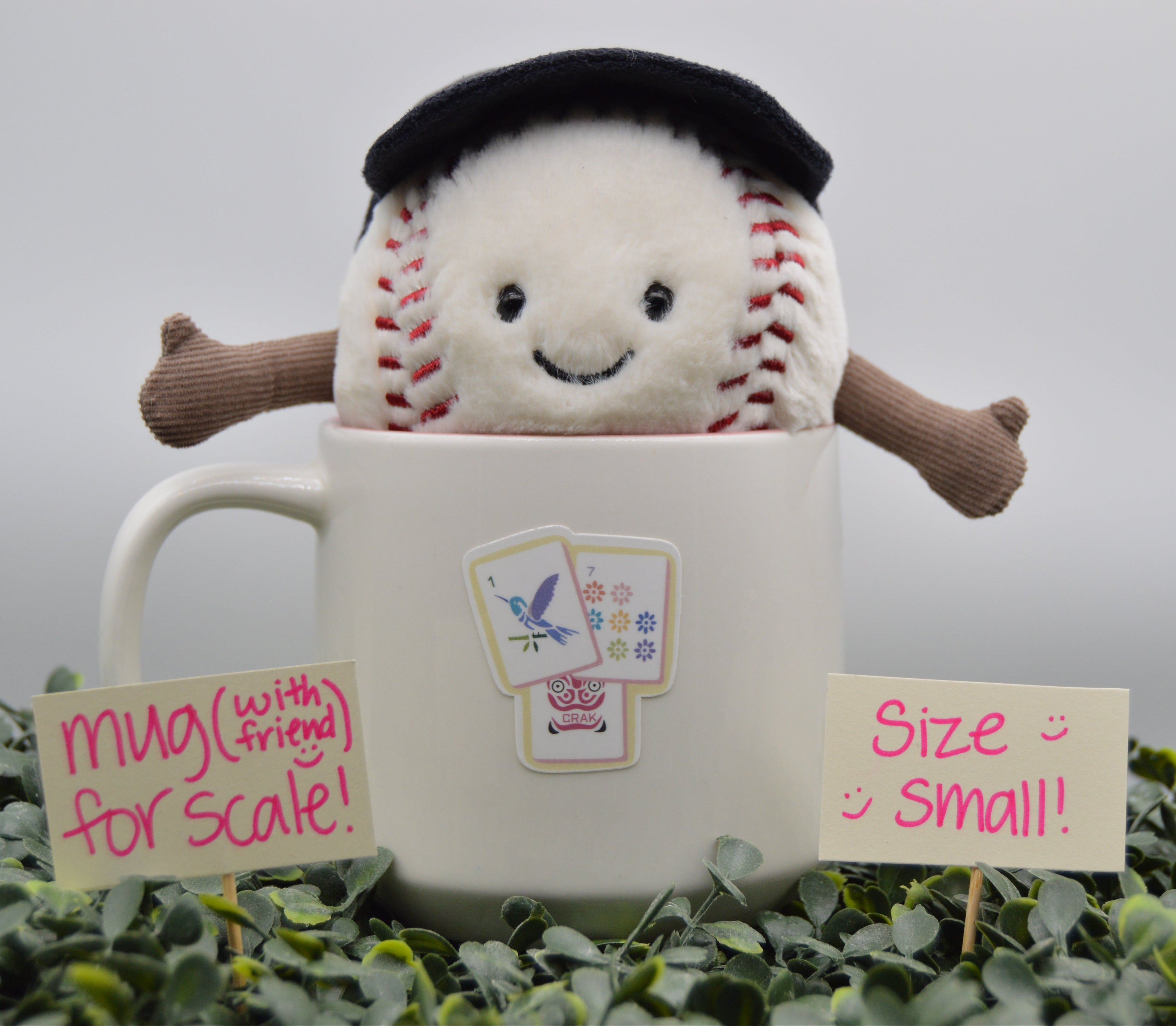 A plush baseball toy with a black beret and brown arms popping out of a white ceramic mug, surrounded by green foliage with handwritten scale signs reading 'mug (with friend) for scale!' and 'Size small!'.