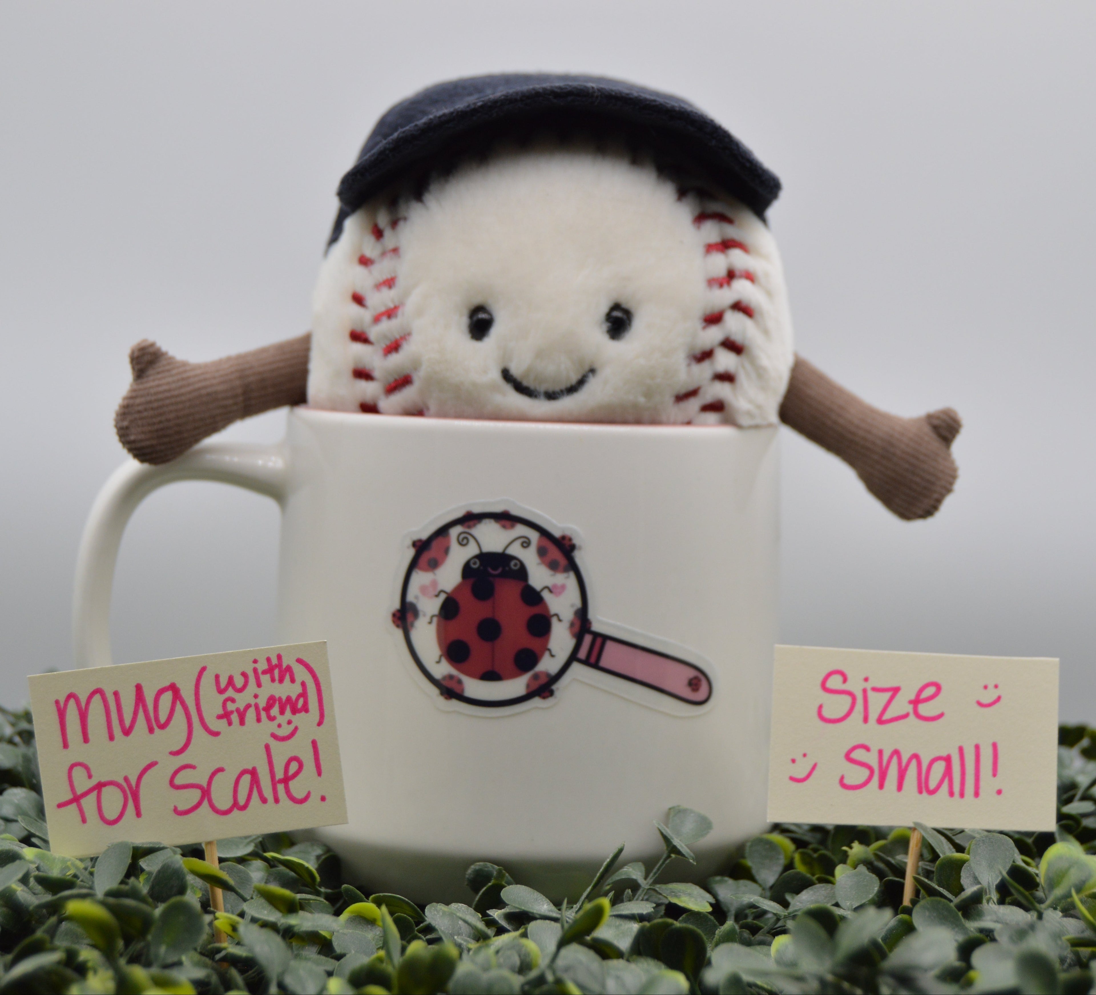 A small plush baseball toy wearing a dark navy cap, peeking out from behind a white ceramic mug filled with green leaves. Two small paper signs read 'mua (with friends) for scale!' and 'Size small!'.