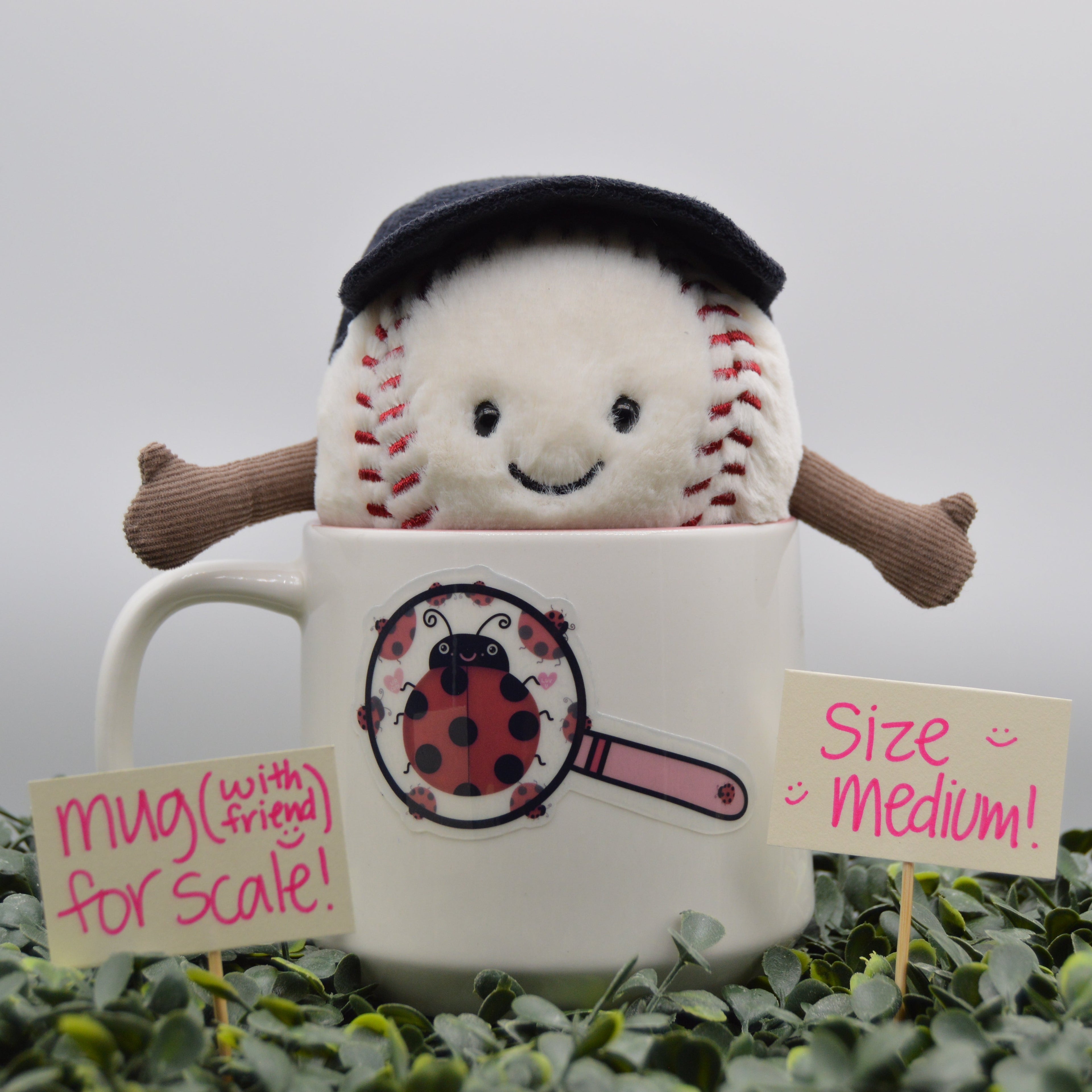 A plush baseball toy with a black cap and brown arms popping out of a white ceramic mug decorated with a ladybug magnifying glass graphic. Small signs reading 'mug (with string for scale!)', 'Size Medium!', and a ladybug sticker are placed in green foliage against a soft gray background.
