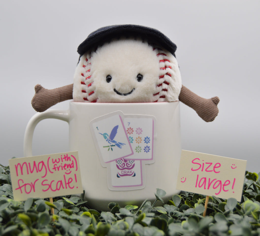 A plush baseball toy wearing a black beret sits inside a large white ceramic mug, surrounded by green foliage and two handwritten scale signs reading 'mug (with friend) for scale!' and 'size large!'.