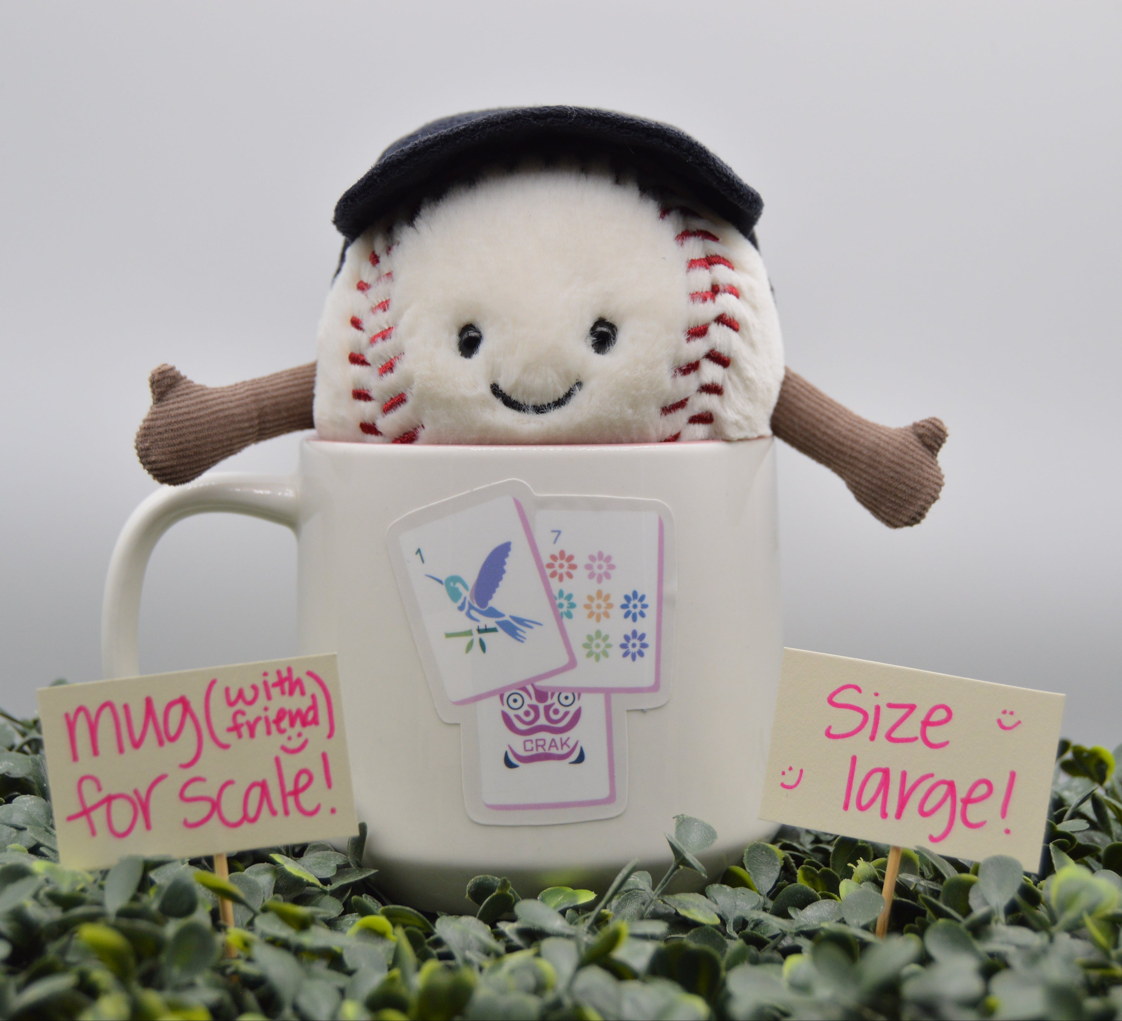 A plush baseball toy wearing a black beret sits inside a large white ceramic mug, surrounded by green foliage and two handwritten scale signs reading 'mug (with friend) for scale!' and 'size large!'.