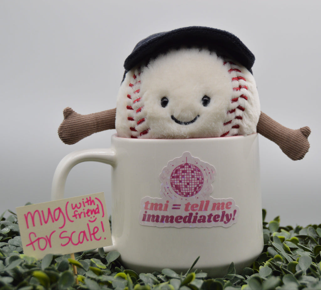 A plush baseball character wearing a black beret sits inside a white ceramic mug with pink text reading 'tmi = tell me immediately!'. The mug is placed on green foliage against a soft gray background, with a small handwritten note that says 'mug (with friend) for scale!'.