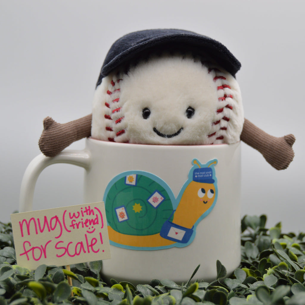 A plush baseball toy with a smiling face, red stitching, and small brown arms peeks out from a white ceramic mug decorated with a colorful snail illustration. The mug sits on green foliage against a soft gray background, with a small sign reading 'mug (with friend) for scale!'.