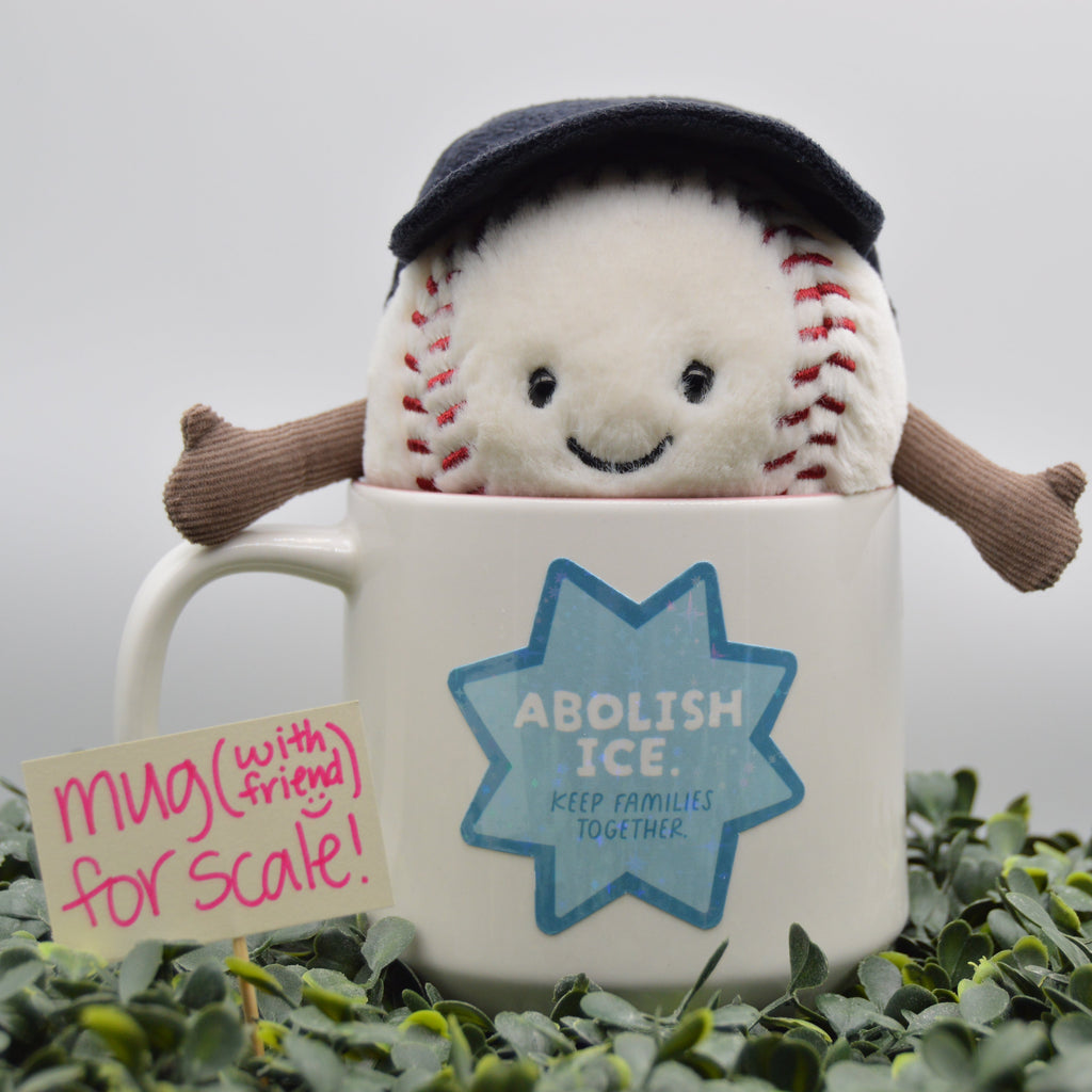 A plush baseball toy wearing a dark navy cap and brown arms, emerging from a white ceramic mug with a blue 'ABOLISH ICE' star graphic. A small sign reading 'mug (with friend) for scale!' sits beside it, and the mug is surrounded by green foliage against a soft gray background.