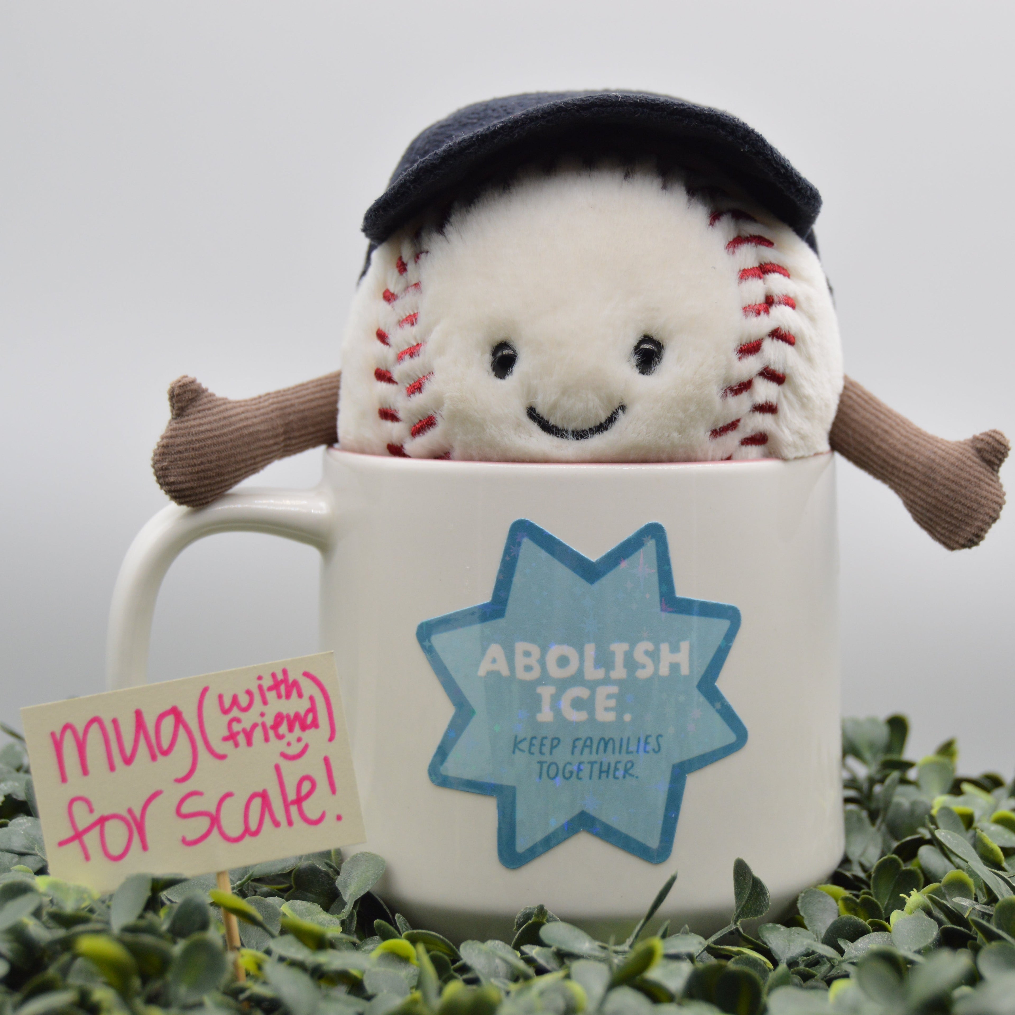 A plush baseball toy wearing a dark navy cap and brown arms, emerging from a white ceramic mug with a blue 'ABOLISH ICE' star graphic. A small sign reading 'mug (with friend) for scale!' sits beside it, and the mug is surrounded by green foliage against a soft gray background.