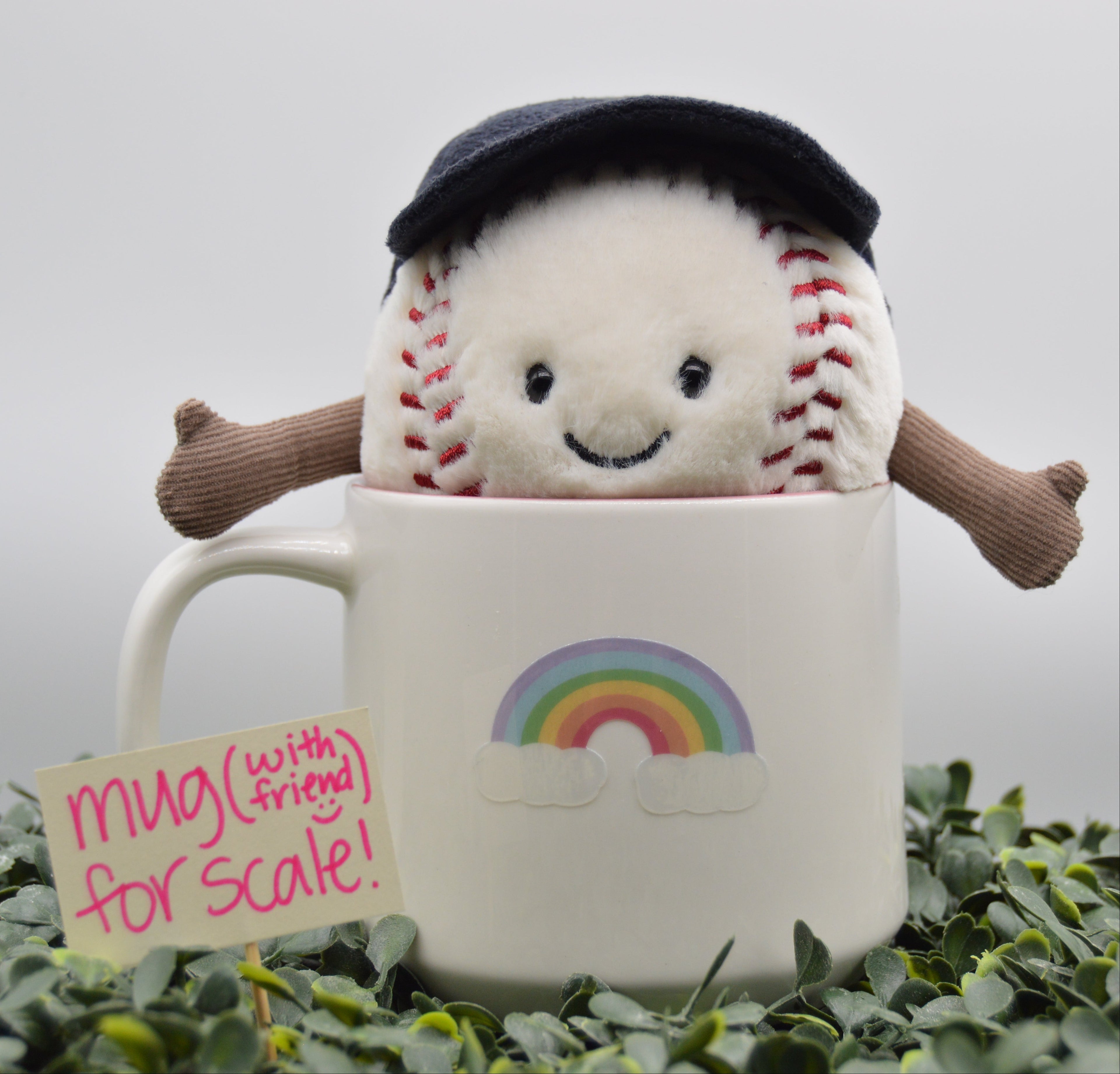 A plush baseball toy with a black beret and brown arms peeks out of a white ceramic mug decorated with a rainbow. The setup is surrounded by green foliage on a light gray background, with a small handwritten note that reads 'mug (with friend) for scale!'.