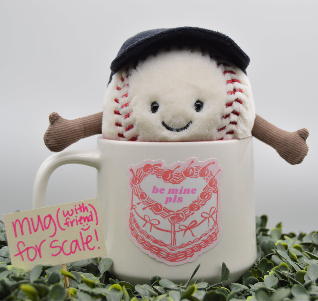 A white ceramic mug with a pink cake graphic and the words 'be mine pie' sits in green foliage, with a plush baseball character peeking out from inside the mug. A small sign next to it reads 'mug (with friend) for scale!'.