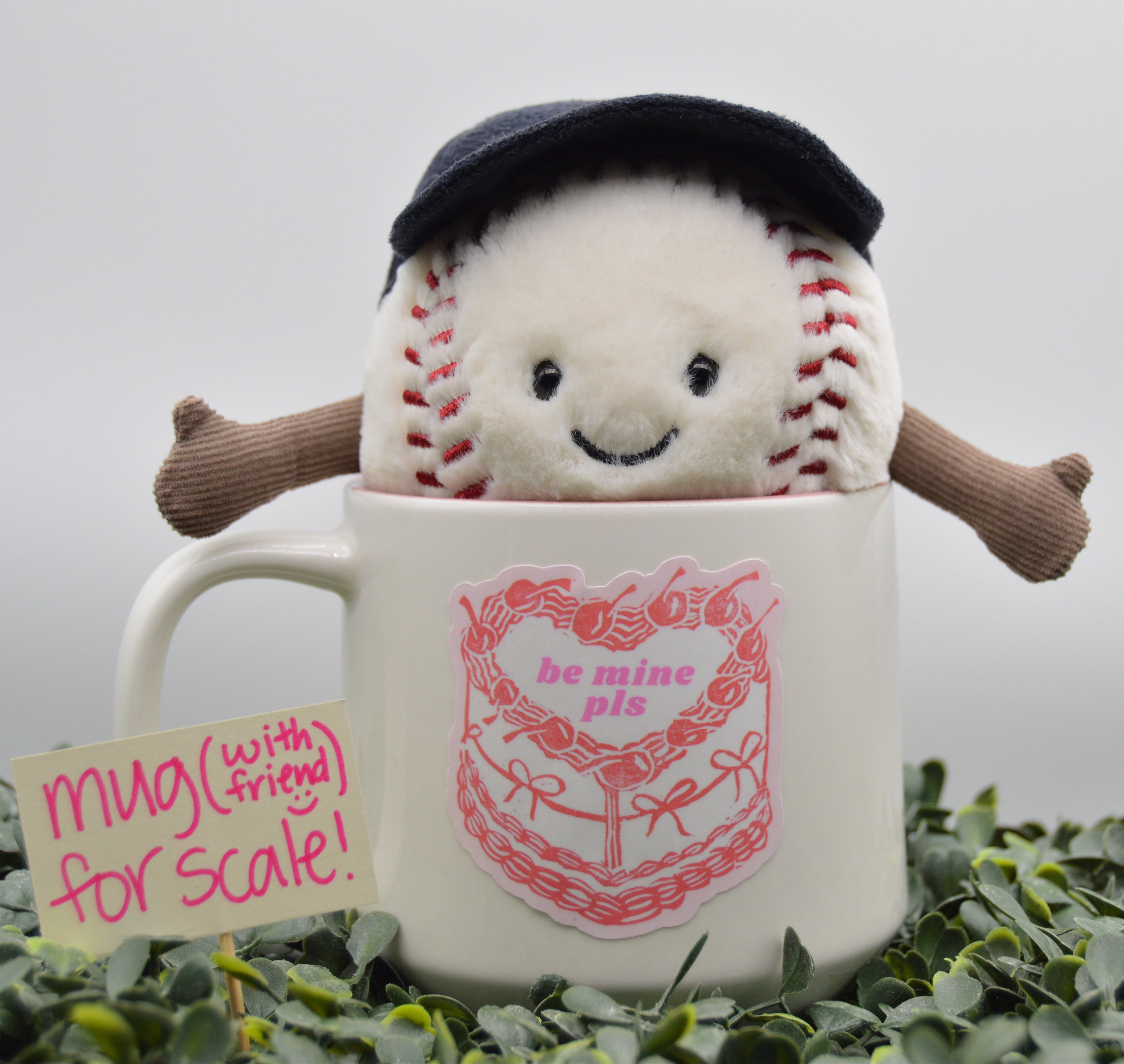 A white ceramic mug with a pink cake graphic and the words 'be mine pie' sits in green foliage, with a plush baseball character peeking out from inside the mug. A small sign next to it reads 'mug (with friend) for scale!'.