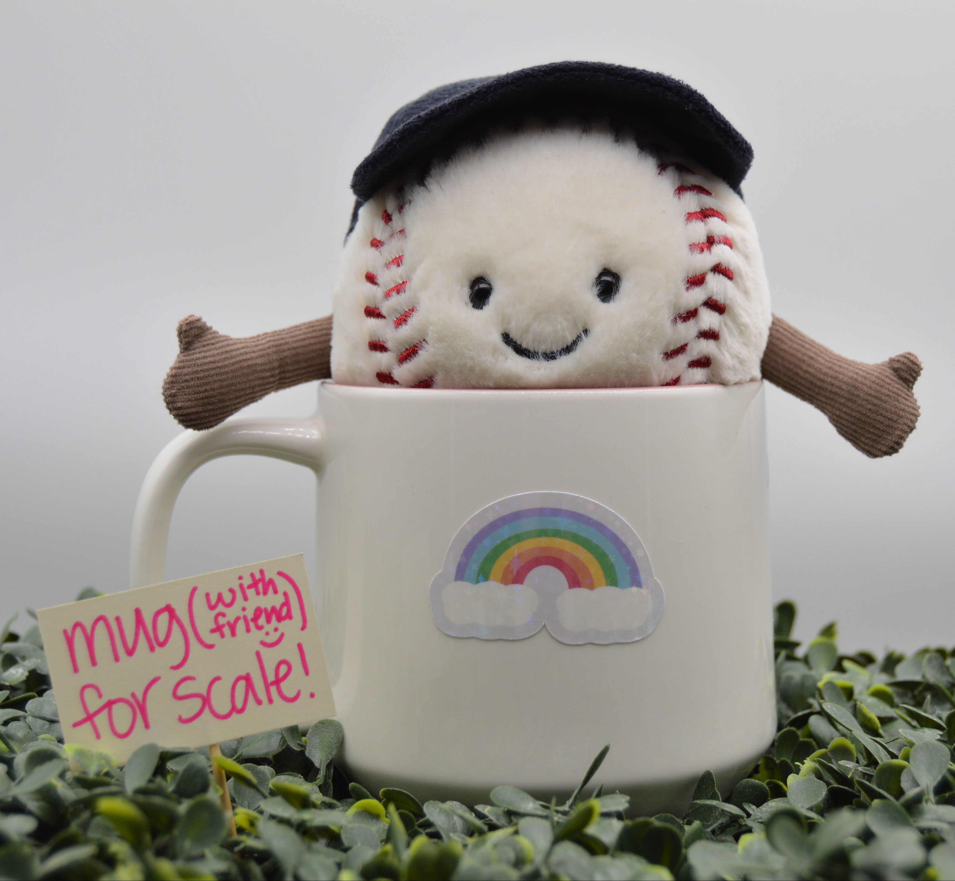 A plush baseball character wearing a black beret sits inside a white ceramic mug with a rainbow sticker. The mug is placed on green foliage against a soft gray background, with a small note that reads 'mug (with fringe) for scale!'.