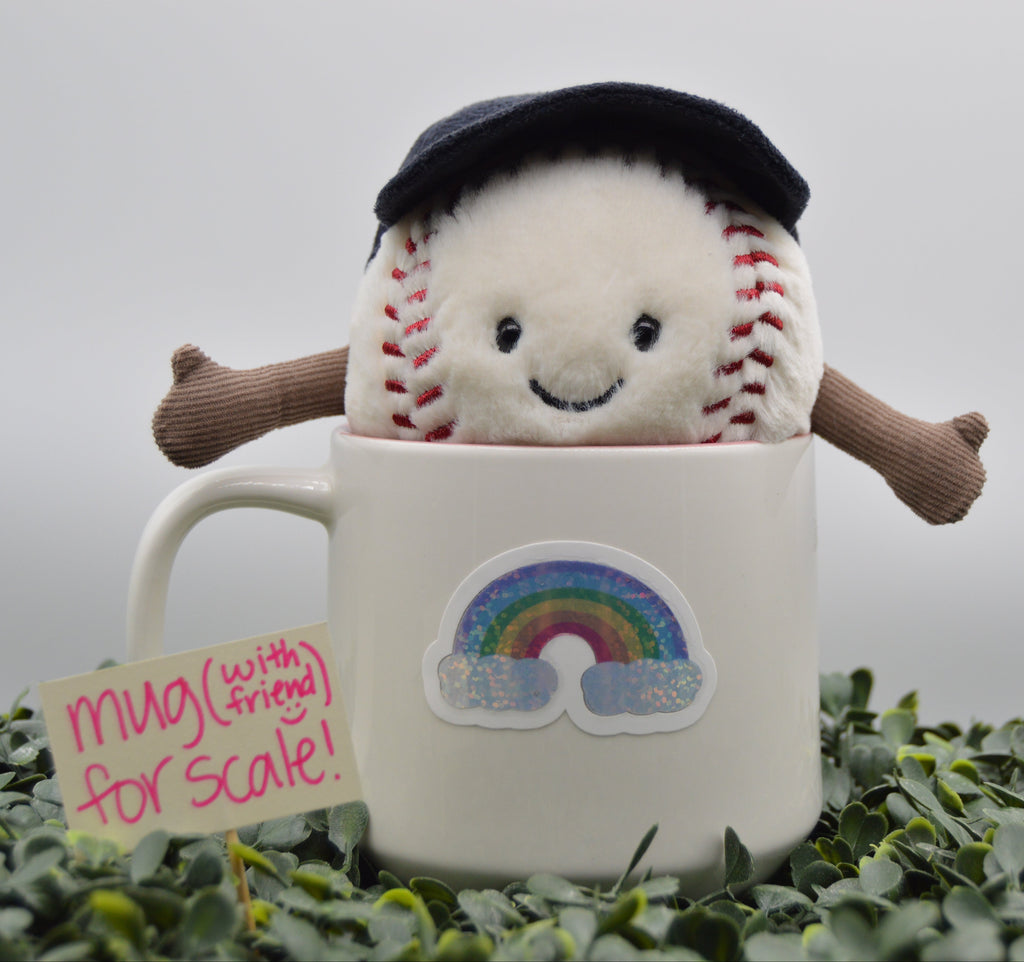 A plush baseball character wearing a black beret and brown arms sits inside a white ceramic mug with a rainbow sticker. A small sign next to the mug reads 'mug (with friend) for scale!'.