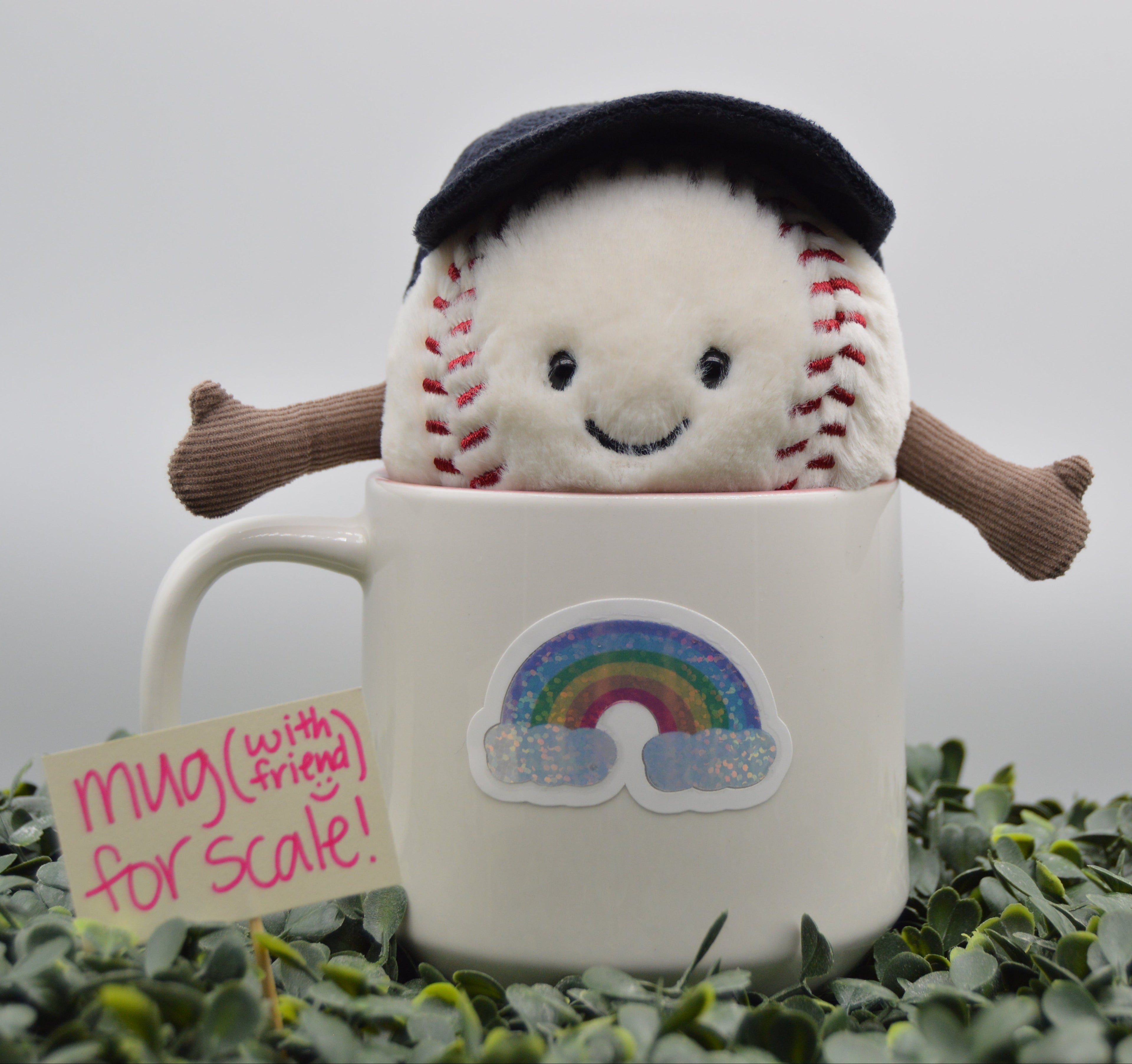 A plush baseball character wearing a black beret and brown arms sits inside a white ceramic mug with a rainbow sticker. A small sign next to the mug reads 'mug (with friend) for scale!'.
