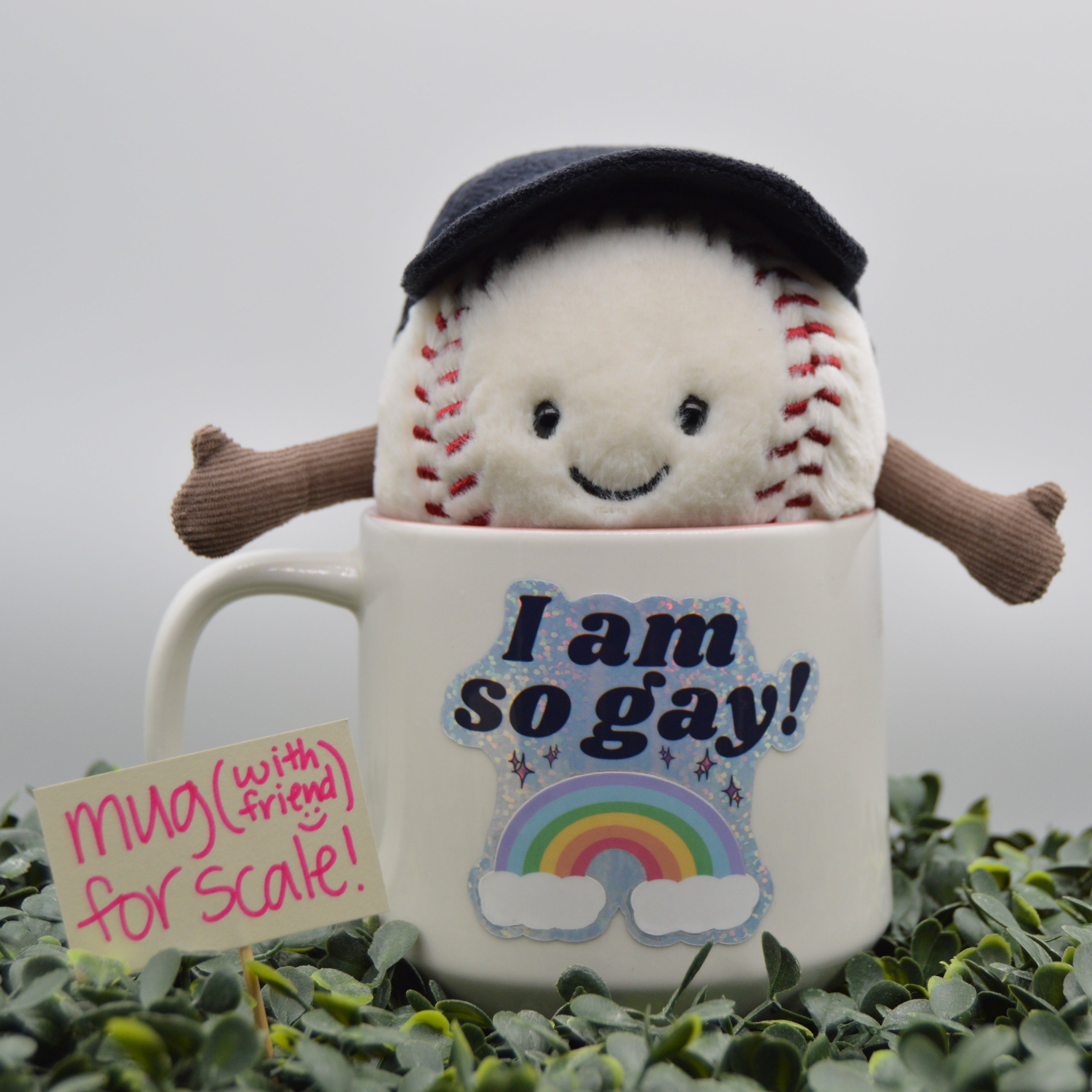 A white ceramic mug with a plush baseball character peeking out from behind it, wearing a dark navy beret and brown arms. The mug features the text 'I am so gay!' above a rainbow graphic.