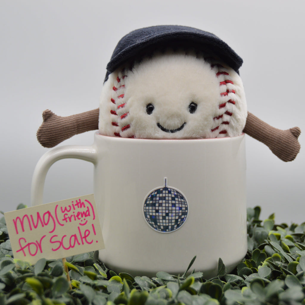 A plush baseball toy with a dark navy blue cap and brown arms, peeking out of a white ceramic mug. The mug has a blue disco ball sticker and a handwritten paper tag that reads 'mua (with friend) for scale!'.