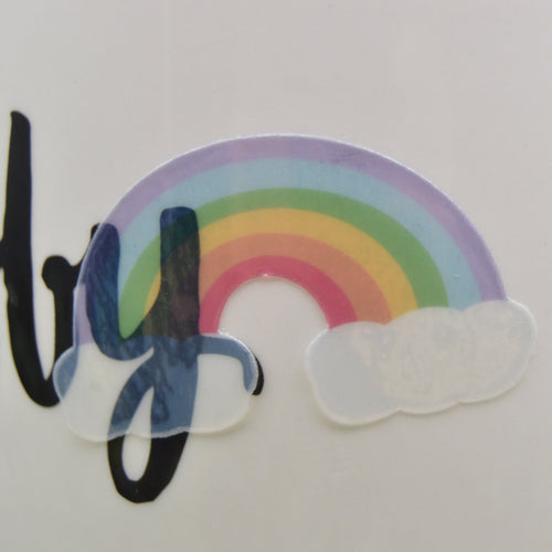 A close-up of a white cylindrical mug or cup featuring a colorful rainbow sticker with white clouds and black script lettering.