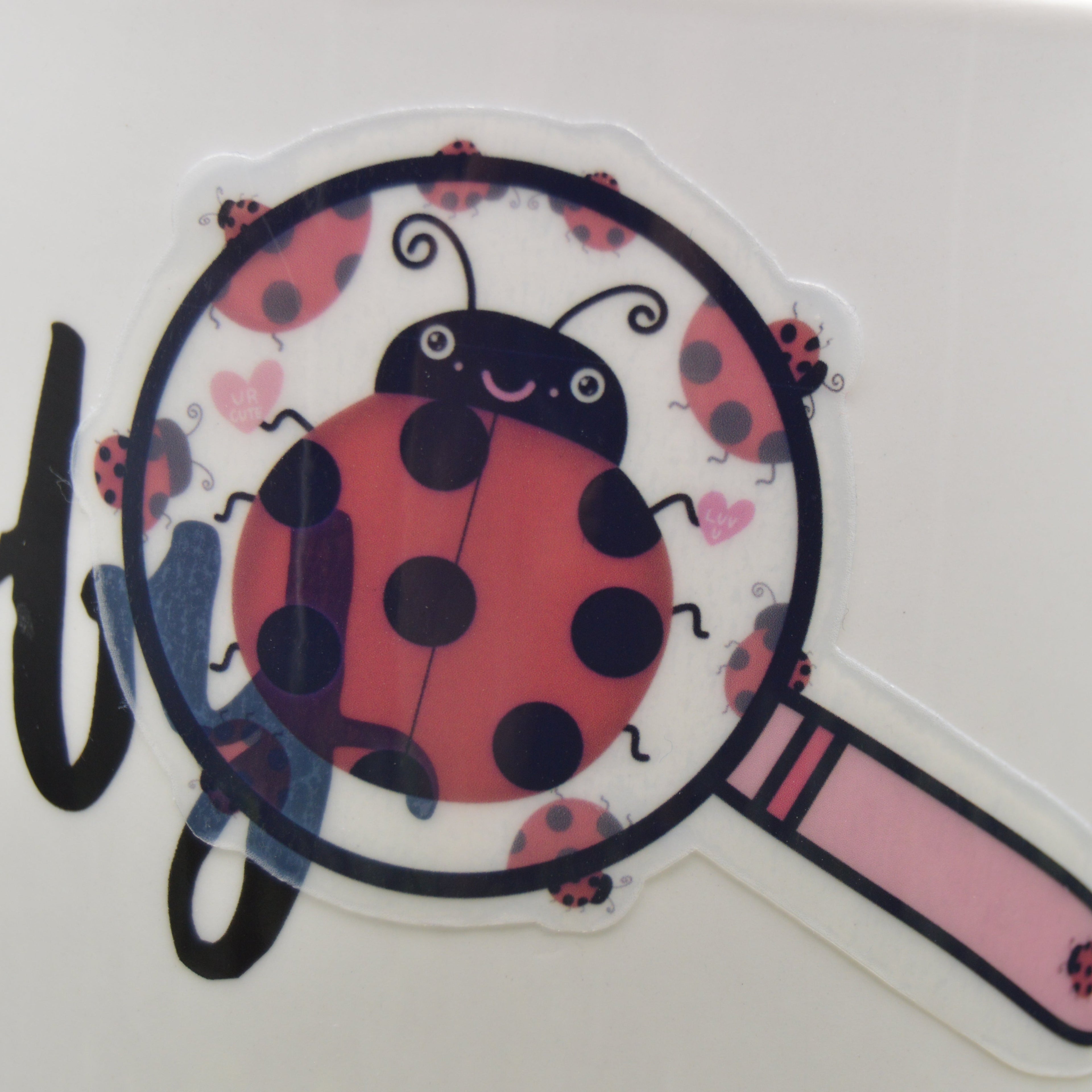 A close-up of a white ceramic mug featuring a cartoon ladybug sticker under a magnifying glass, with partial black script lettering on the left.