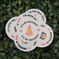 A set of round white paper party stickers with birthday-themed text and illustrations, arranged on a background of green leafy foliage.