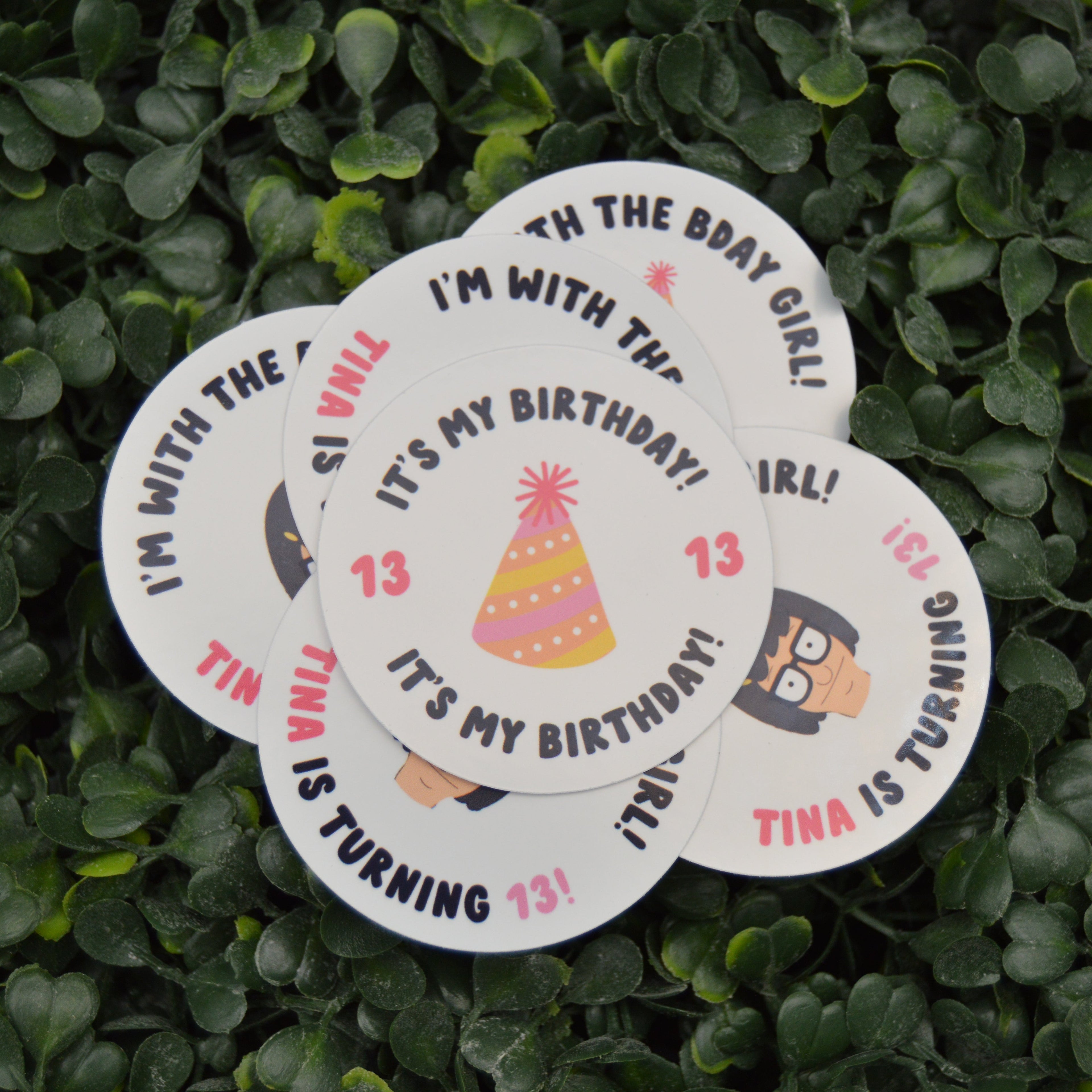 A set of round white paper party stickers with birthday-themed text and illustrations, arranged on a background of green leafy foliage.