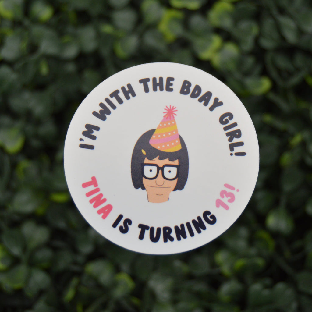 A round white sticker with a cartoon illustration of a girl with glasses and a party hat, surrounded by the text 'I'M WITH THE BDAY GIRL! TINA IS TURNING 13!' in black and pink lettering, shown against a blurred green hedge background.