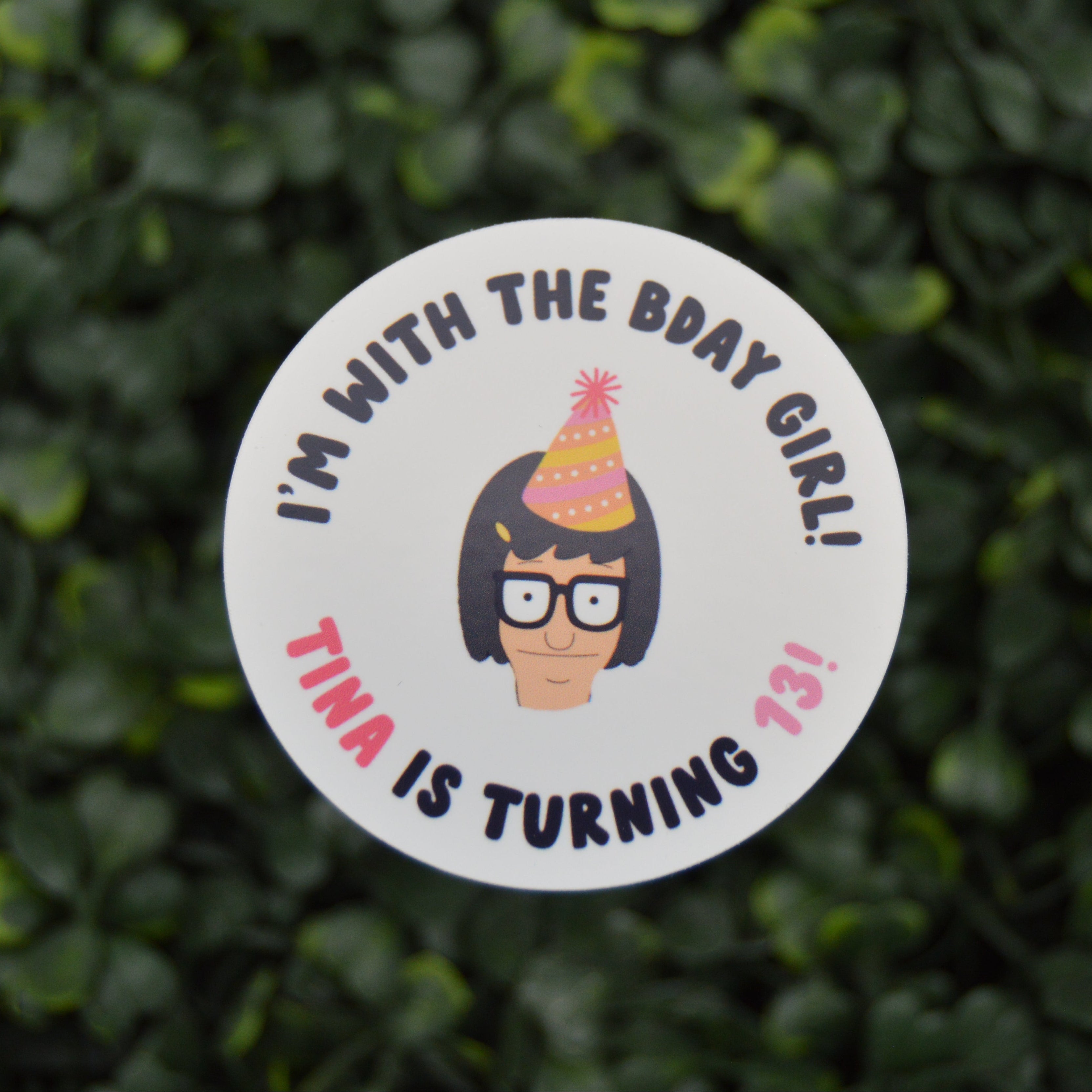 A round white sticker with a cartoon illustration of a girl with glasses and a party hat, surrounded by the text 'I'M WITH THE BDAY GIRL! TINA IS TURNING 13!' in black and pink lettering, shown against a blurred green hedge background.