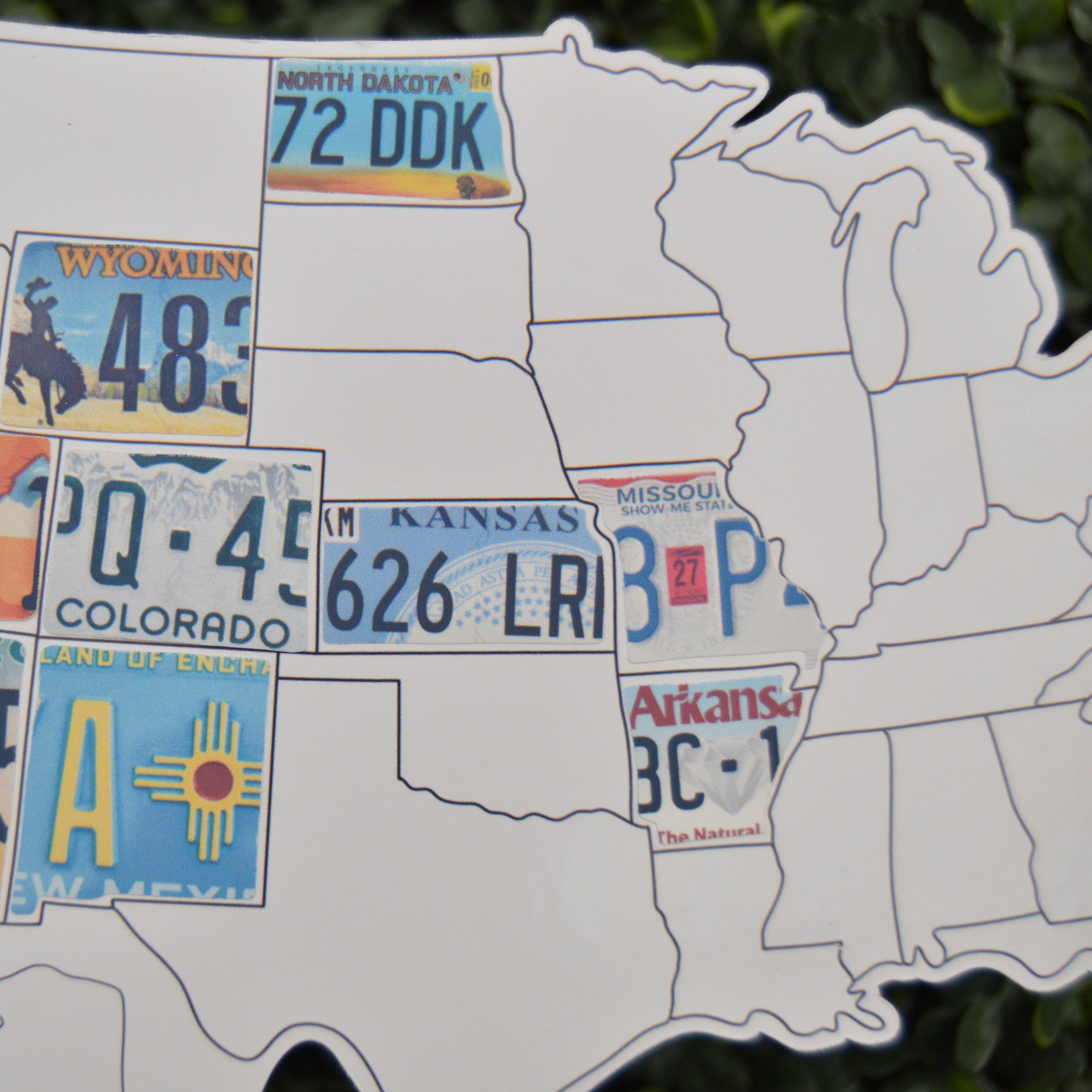 A white cutout map of the United States decorated with colorful vintage license plates, photographed against a blurred green outdoor background.