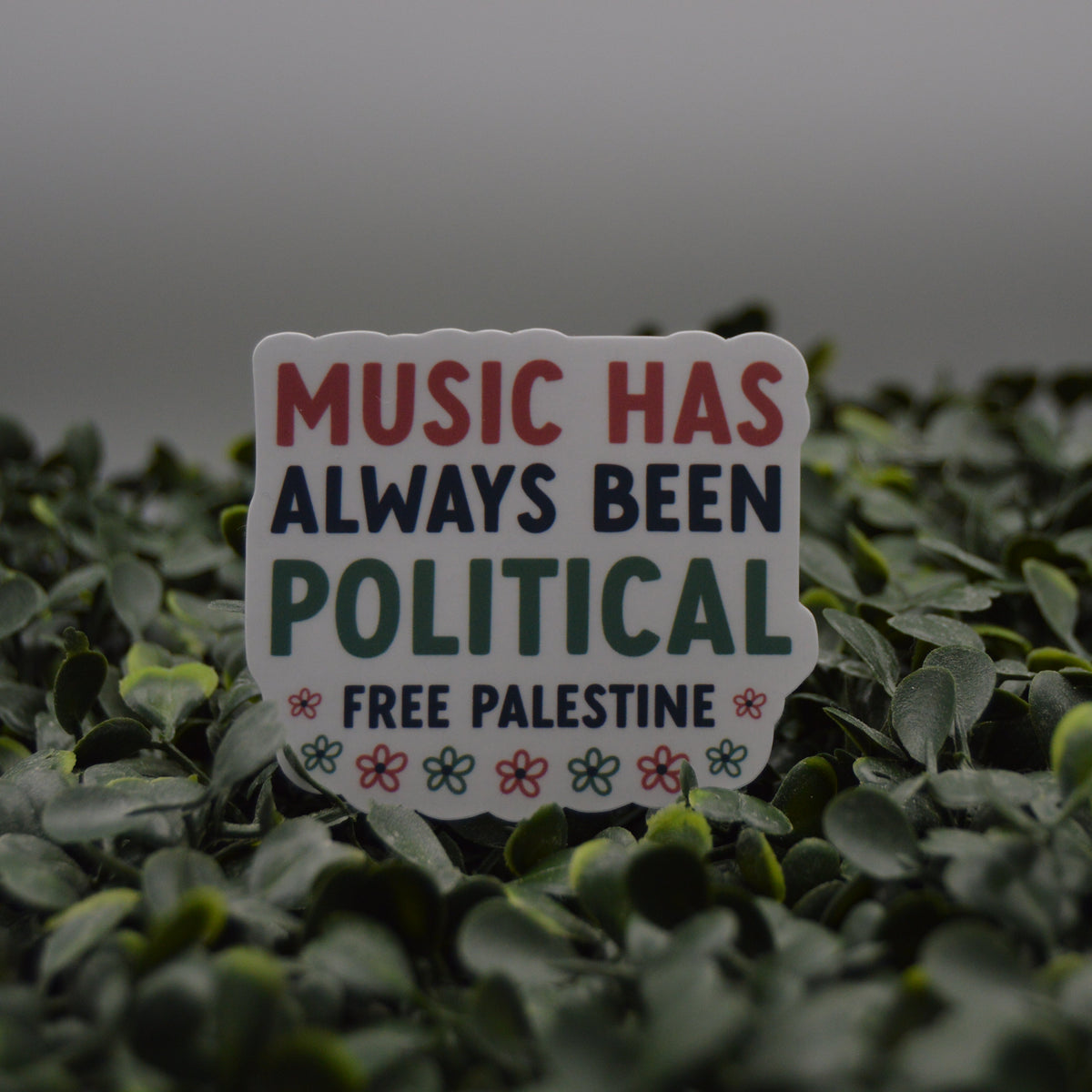 A die-cut sticker with the text 'MUSIC HAS ALWAYS BEEN POLITICAL FREE PALESTINE' in red, navy, and green lettering, placed on a bed of green leaves.