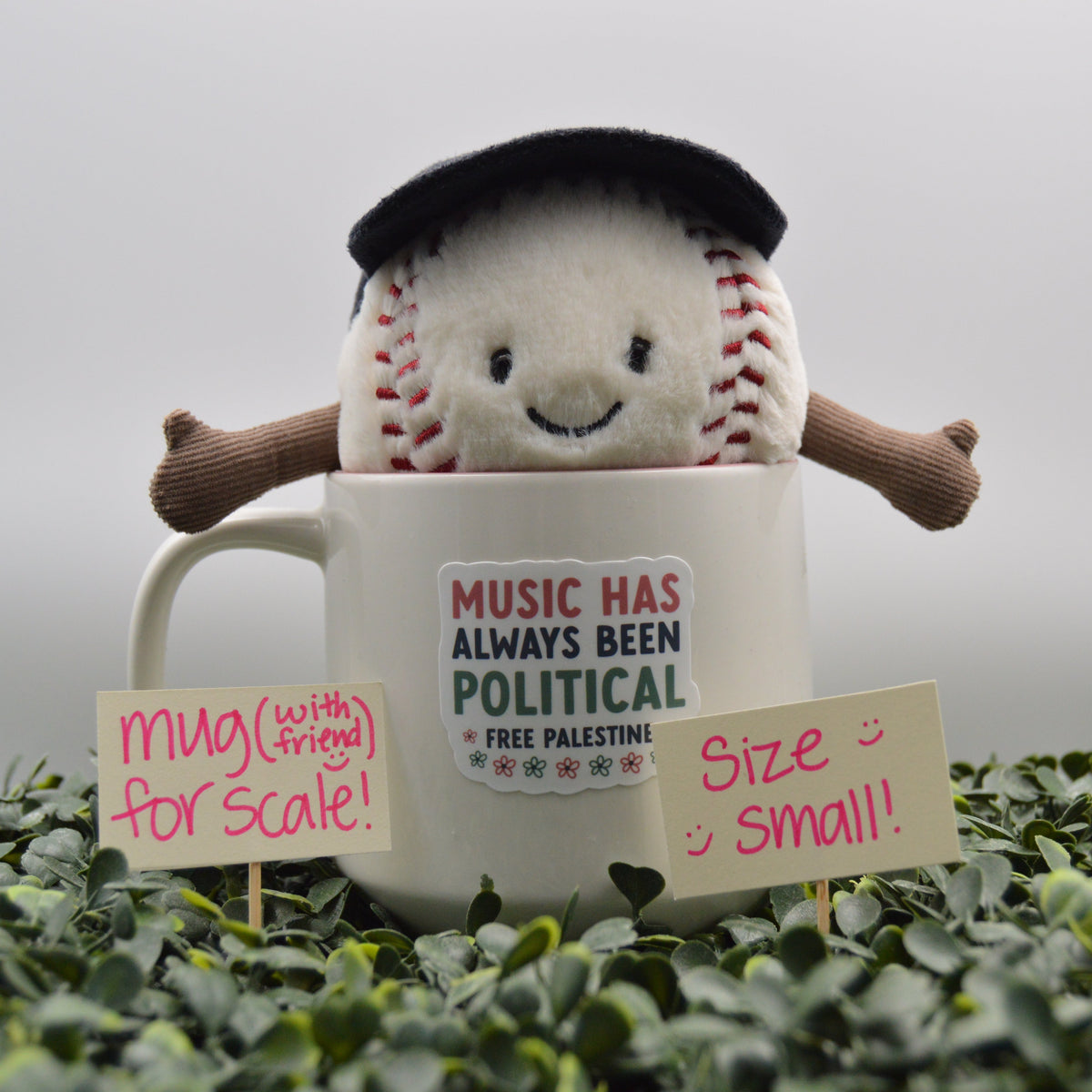 A small plush toy shaped like a baseball with a black beret and brown arms, emerging from a white ceramic mug printed with the text 'MUSIC HAS ALWAYS BEEN POLITICAL FREE PALESTINE'. The scene is placed on green foliage with two handwritten signs reading 'Mug (with friend) for scale!' and 'Size Small!'.