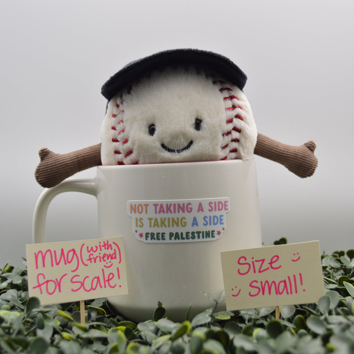 A plush toy baseball wearing a black graduation cap sits inside a white ceramic mug with the text 'NOT TAKING A SIDE IS TAKING A SIDE * FREE PALESTINE'. The mug is surrounded by green foliage and two small signs reading 'mug (with friend) for scale!' and 'Size: small!'.