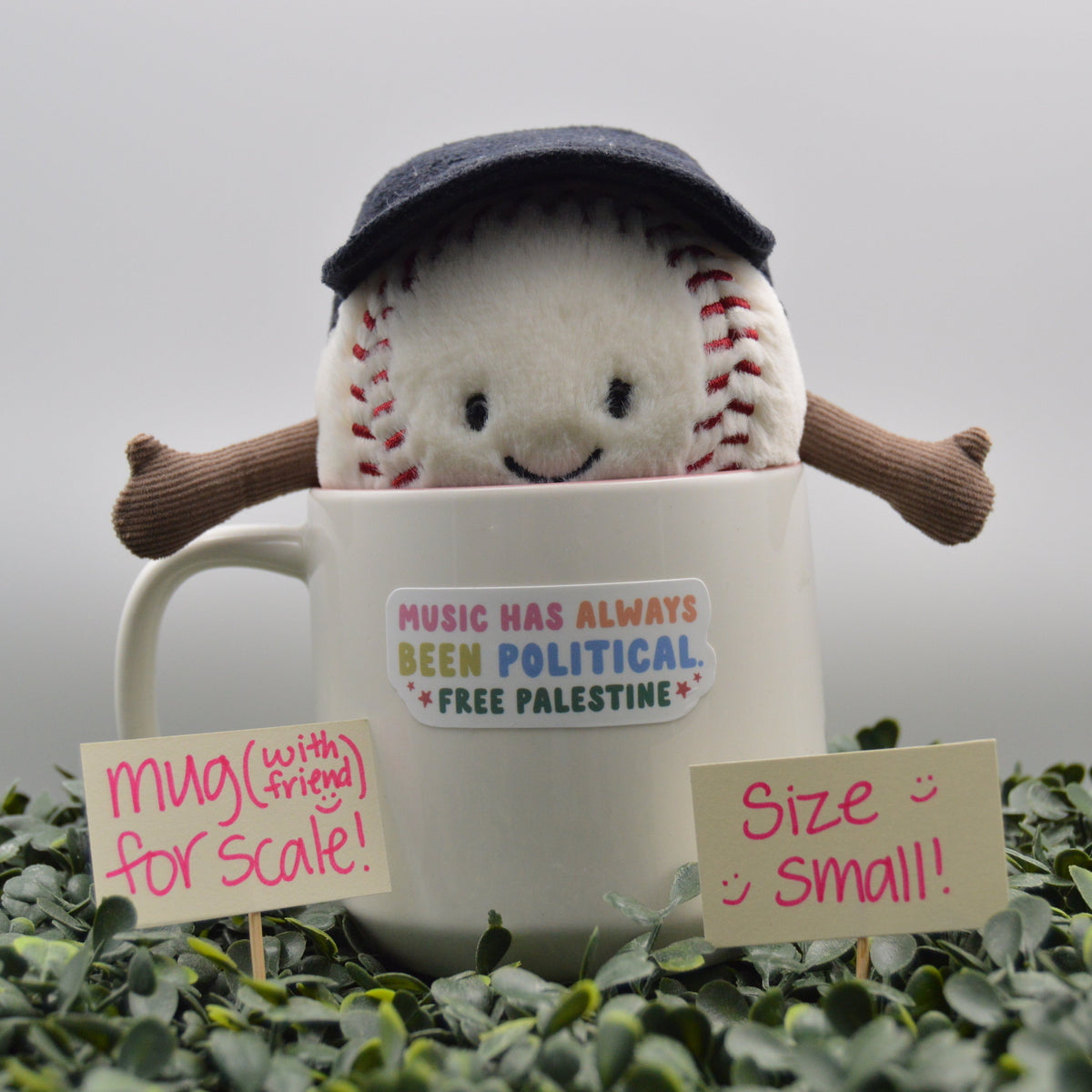 A plush baseball character wearing a dark blue cap and brown arms, sitting inside a white ceramic mug. The mug has a sticker that reads 'MUSIC HAS ALWAYS BEEN POLITICAL - FREE PALESTINE'.