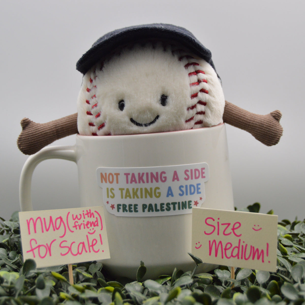 A plush baseball toy wearing a dark navy cap sits inside a white ceramic mug with a printed message. The mug is surrounded by green leaves, with two small paper size and scale signs in front.