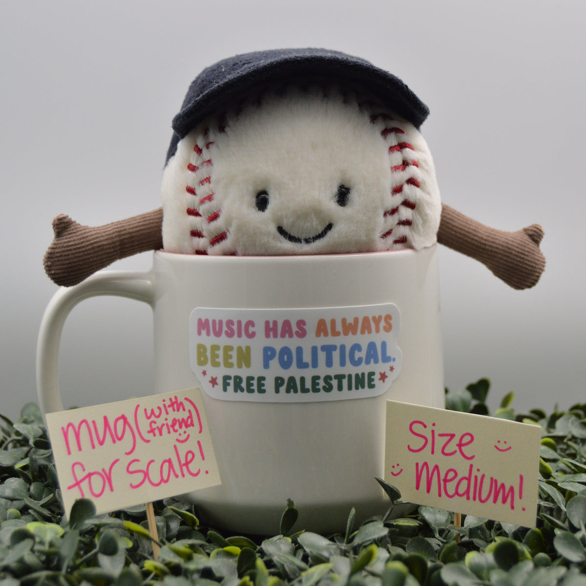A plush baseball toy wearing a dark navy cap sits inside a white ceramic mug with the text 'MUSIC HAS ALWAYS BEEN POLITICAL. FREE PALESTINE.