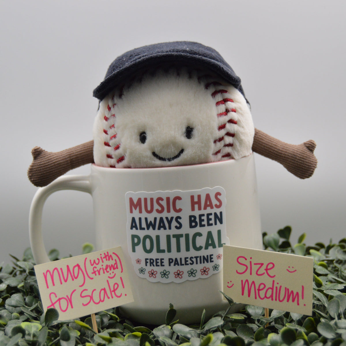 A plush stuffed baseball toy wearing a dark navy cap and brown arms, sitting inside a white ceramic mug with a printed sign that reads 'MUSIC HAS ALWAYS BEEN POLITICAL FREE PALESTINE.' Small handwritten signs say 'mug (with friends) for scale!' and 'size medium!' The scene is styled with green foliage against a soft gray background.
