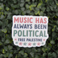 A die-cut sticker with the text 'MUSIC HAS ALWAYS BEEN POLITICAL FREE PALESTINE' in pink, navy, and green lettering, decorated with small flowers, shown against a background of green foliage.