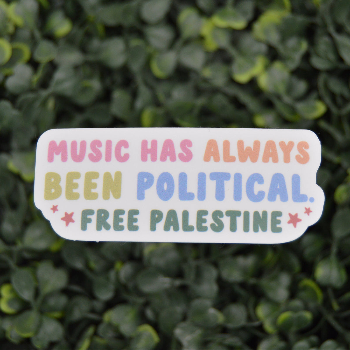 A die-cut sticker with the text 'MUSIC HAS ALWAYS BEEN POLITICAL. FREE PALESTINE' in pastel rainbow colors on a white background, placed against a backdrop of green ivy leaves.