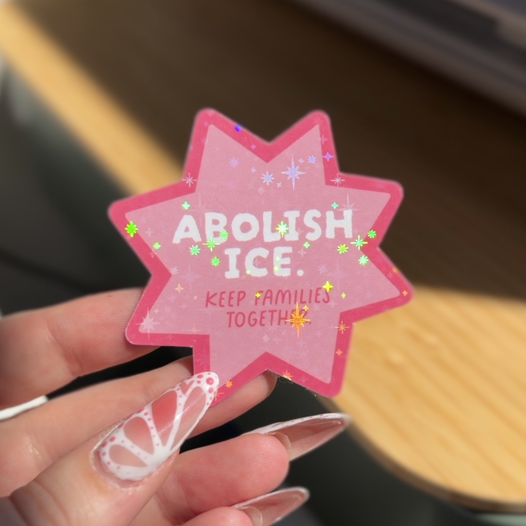 A hand with long white manicured nails holds a pink star-shaped sticker that reads 'ABOLISH ICE. KEEP FAMILIES TOGETHER' in white and yellow text, with small green and yellow floral accents.